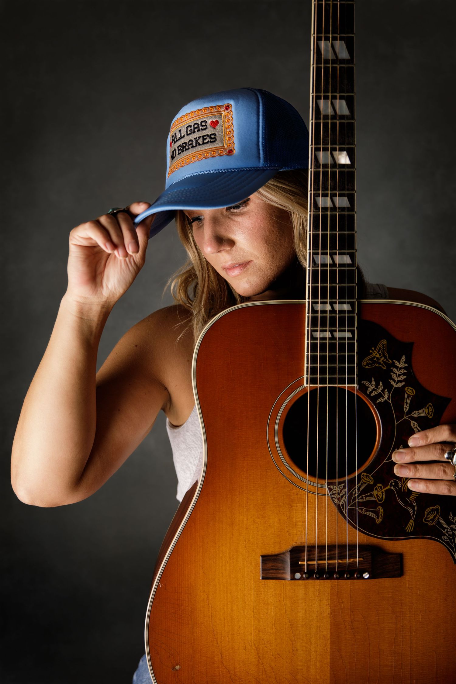 Blonde woman in blue trucker hat poses with acoustic guitar in moody studio portrait.