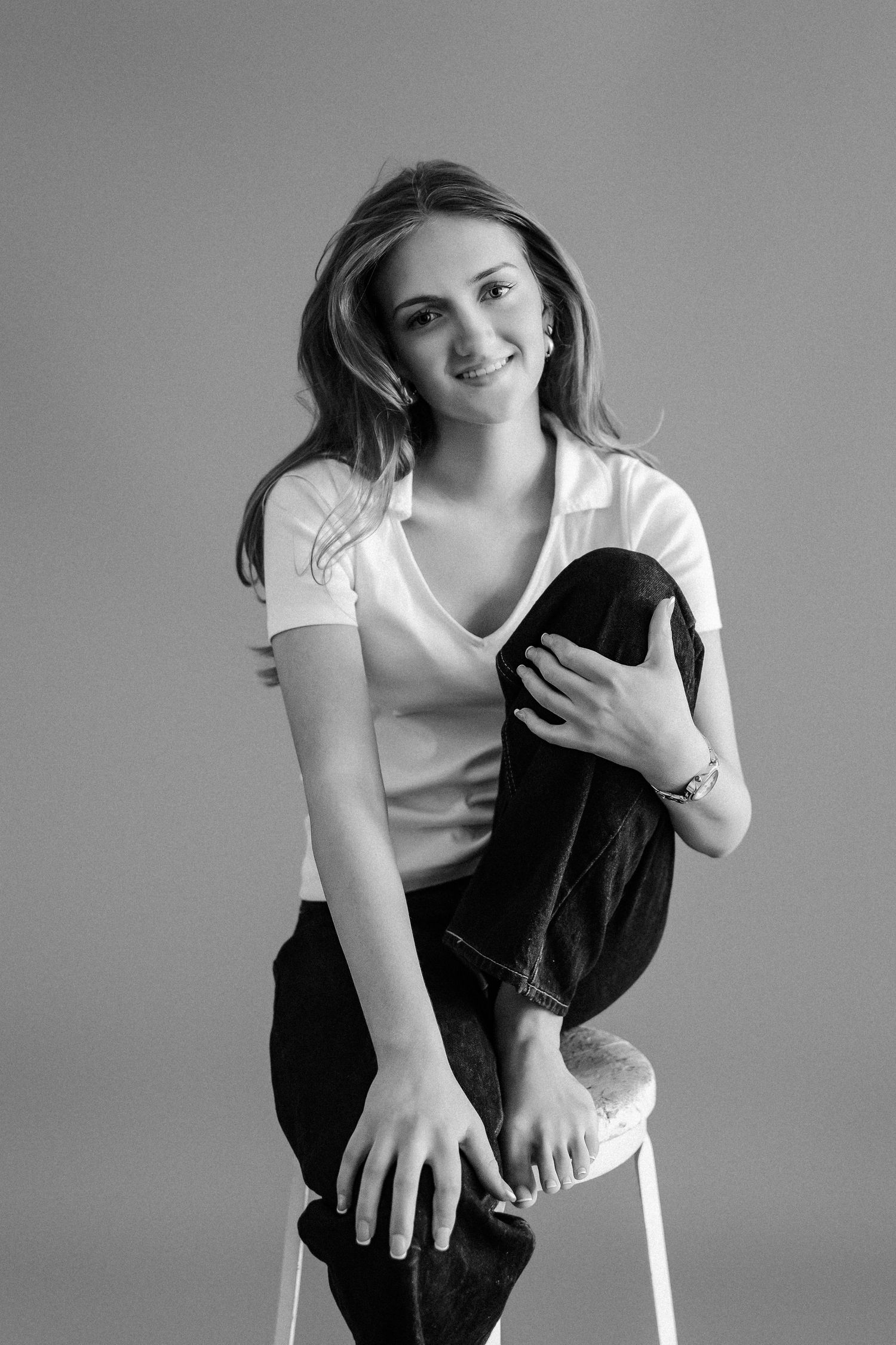 Smiling young woman perched on white stool in white t-shirt and dark jeans, black and white.