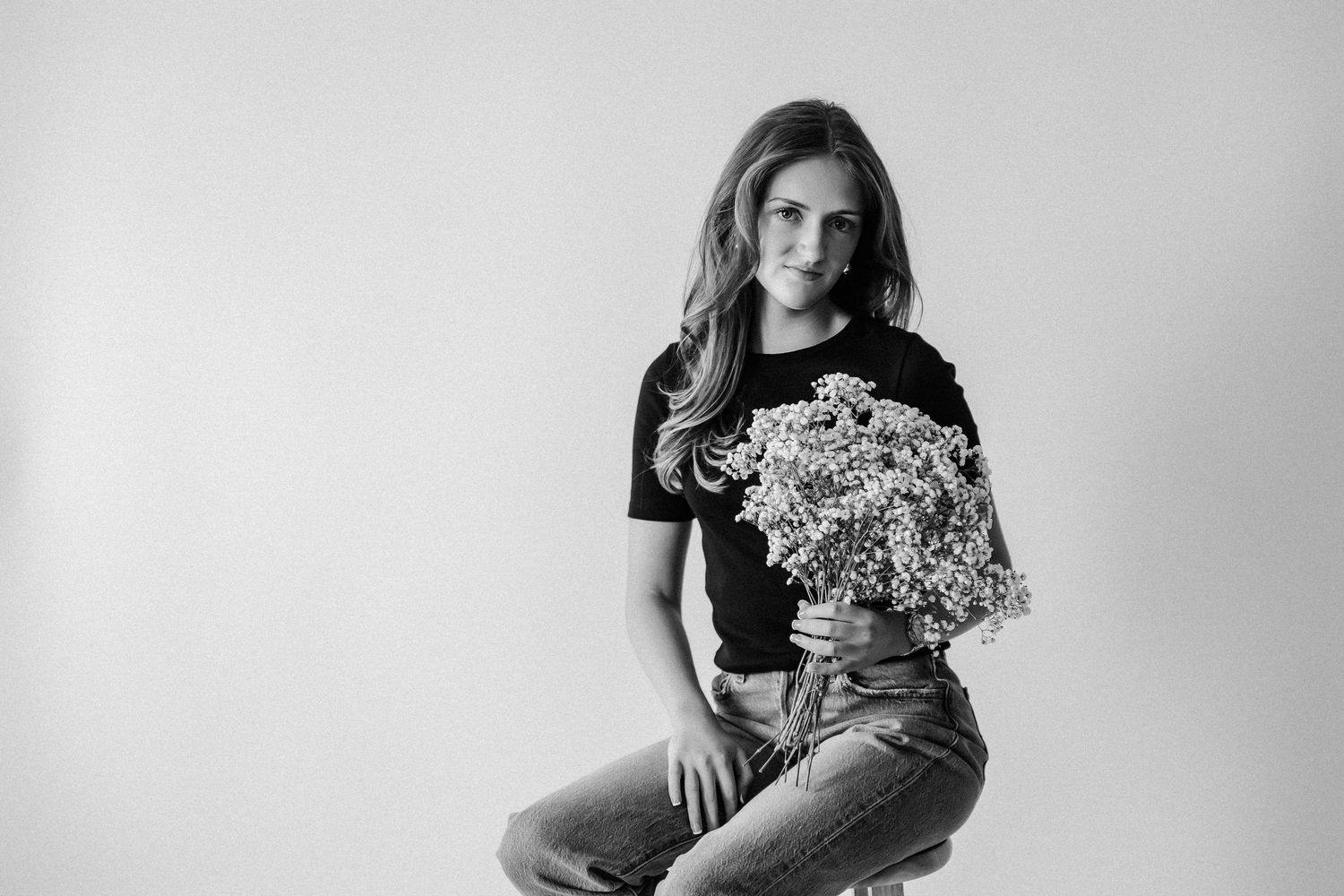 Young woman in black tee and jeans holding white flower bouquet, black and white studio portrait photo.