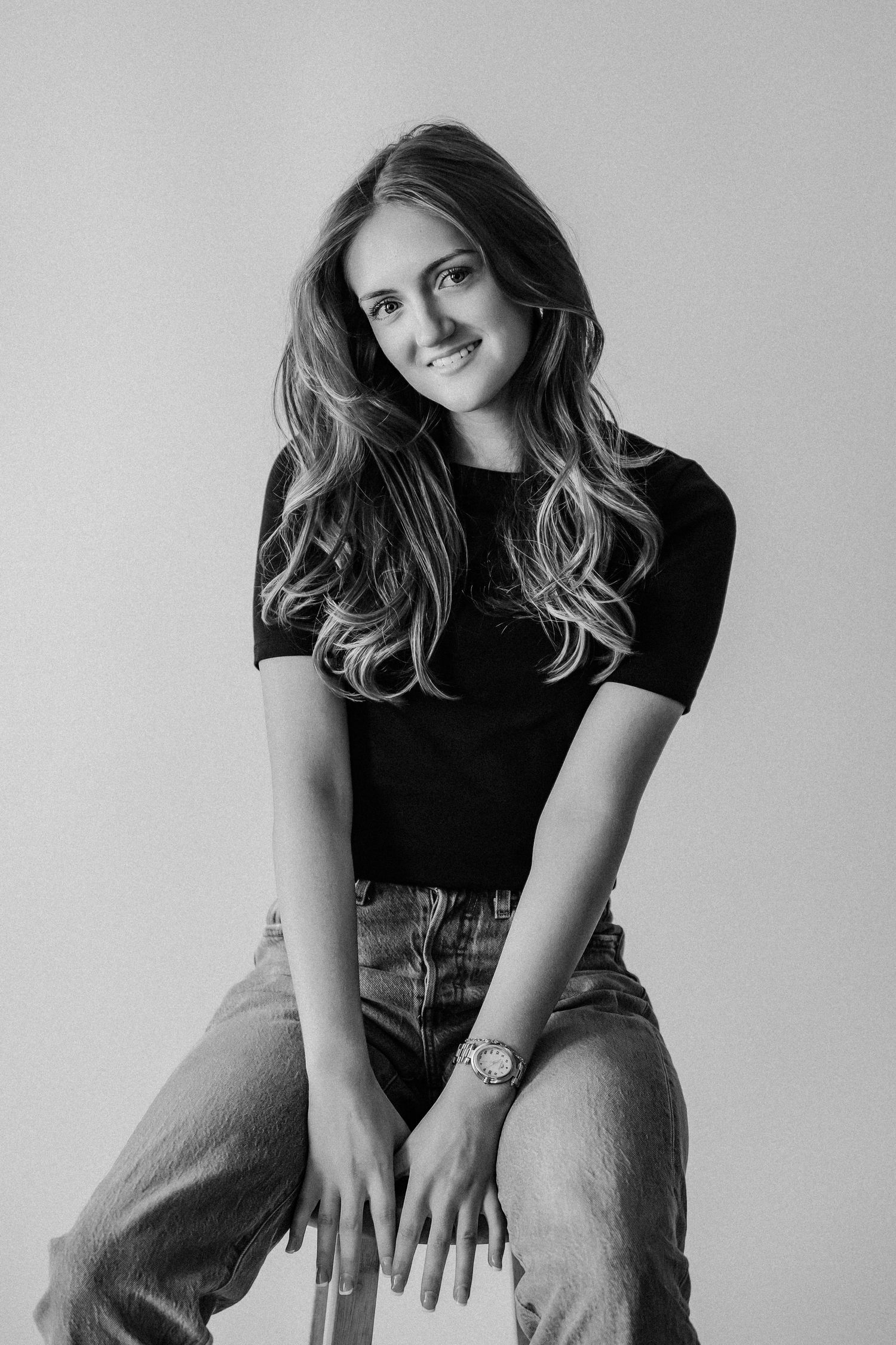 Young woman with wavy hair smiling in black t-shirt and jeans, seated in black and white studio portrait.