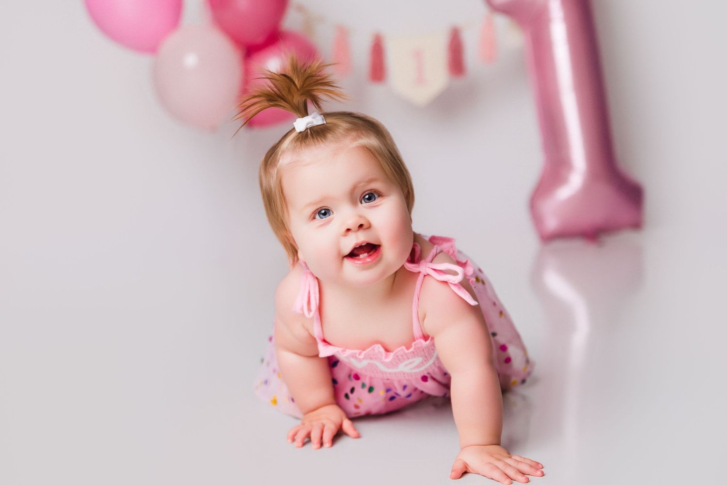 Baby in pink dress posing with pink balloons and party decorations for a first birthday celebration.