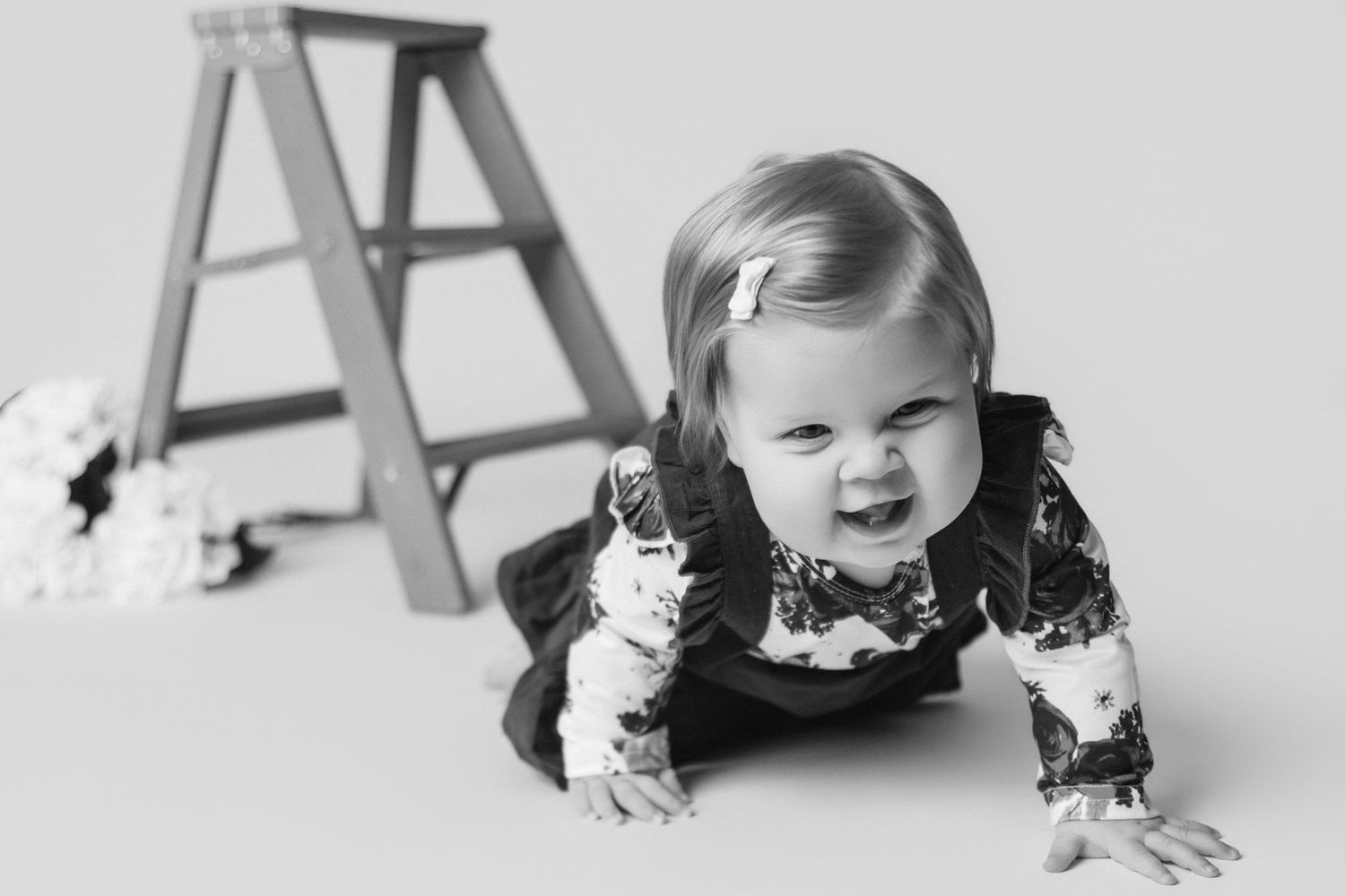 Black and white photo of a smiling baby crawling in floral print outfit near wooden ladder.