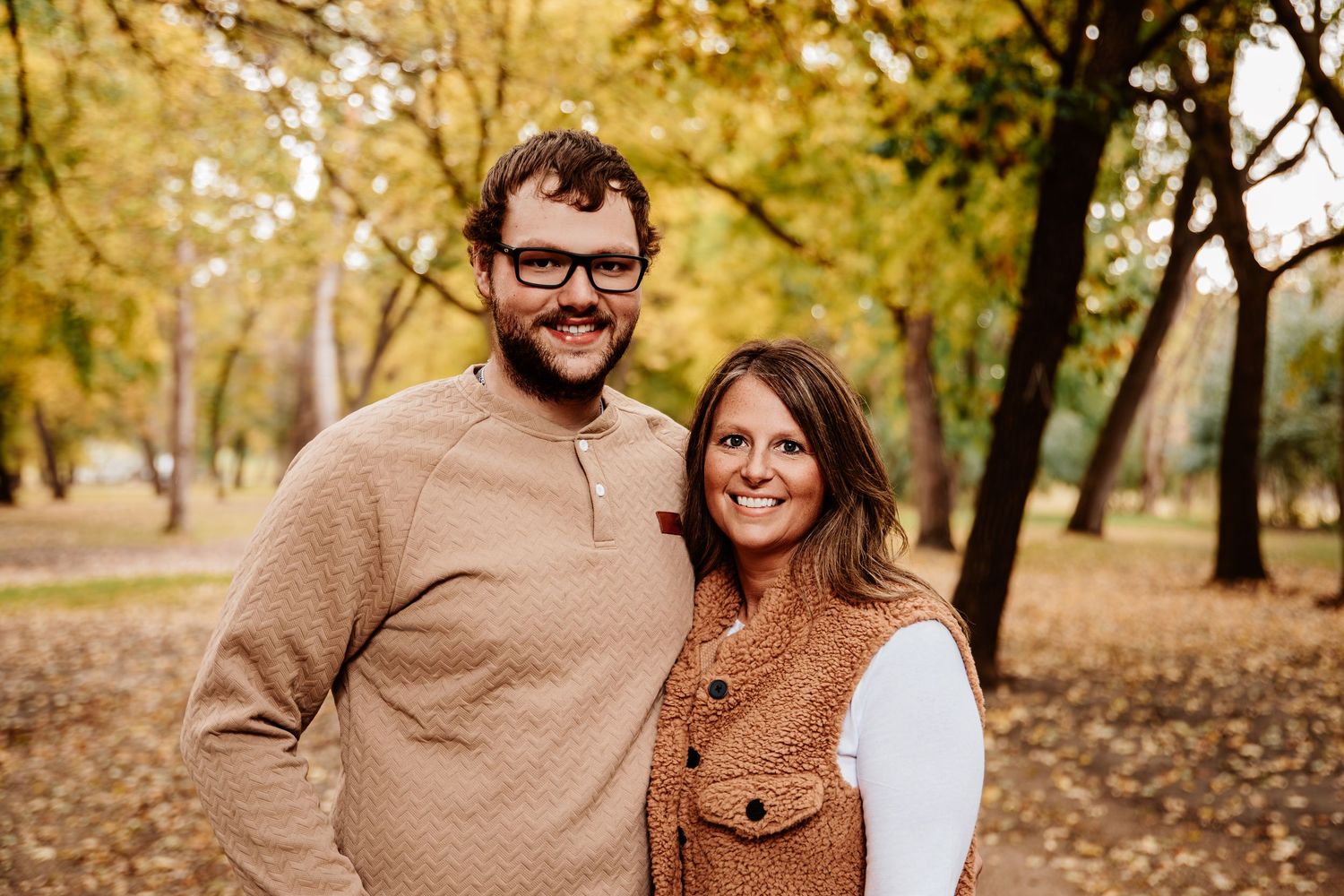 A couple poses together in autumn attire against a background of golden fall foliage.