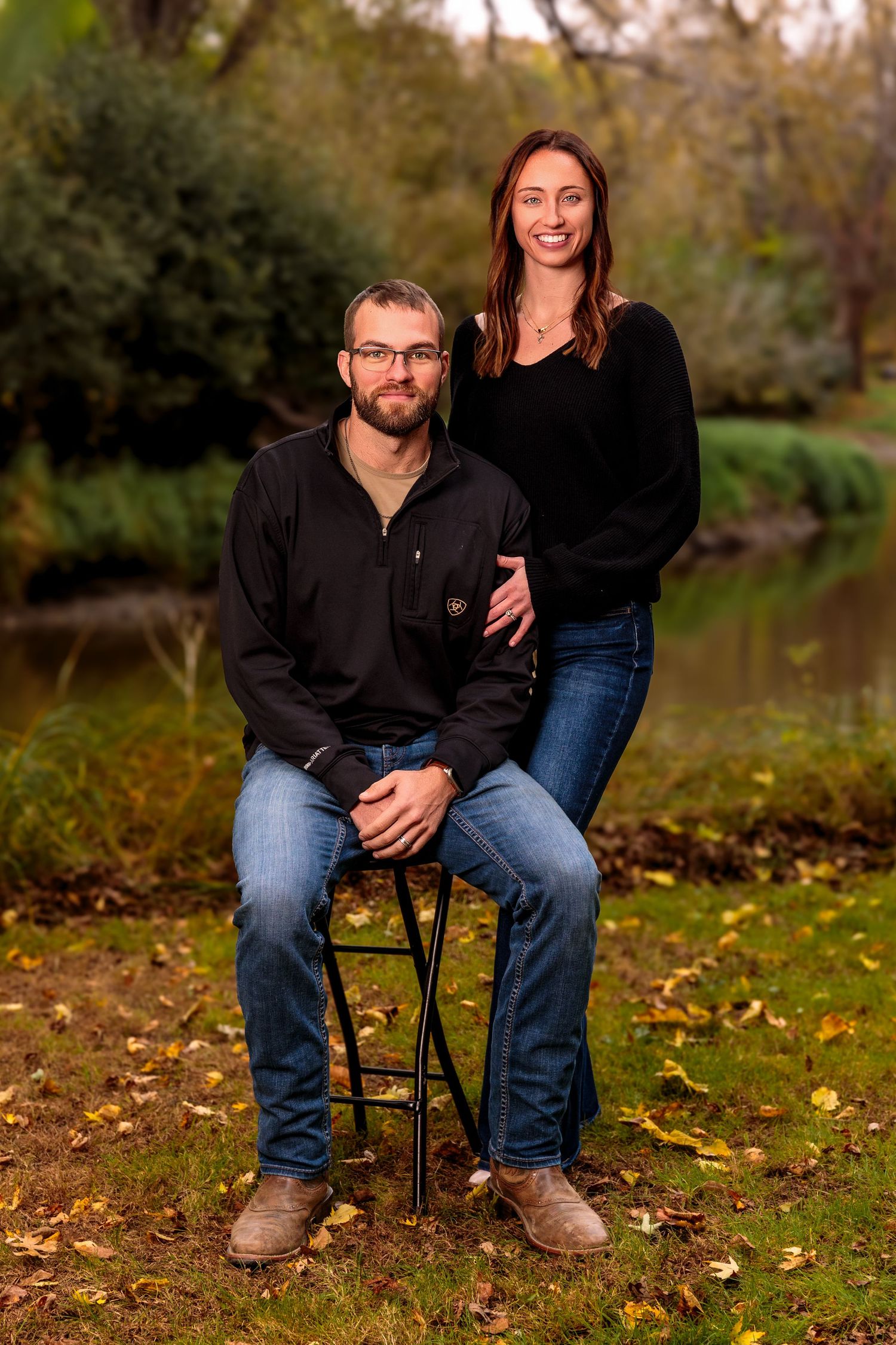 Couple sitting on a chair in an autumn outdoor setting with fallen leaves and a pond in the background.