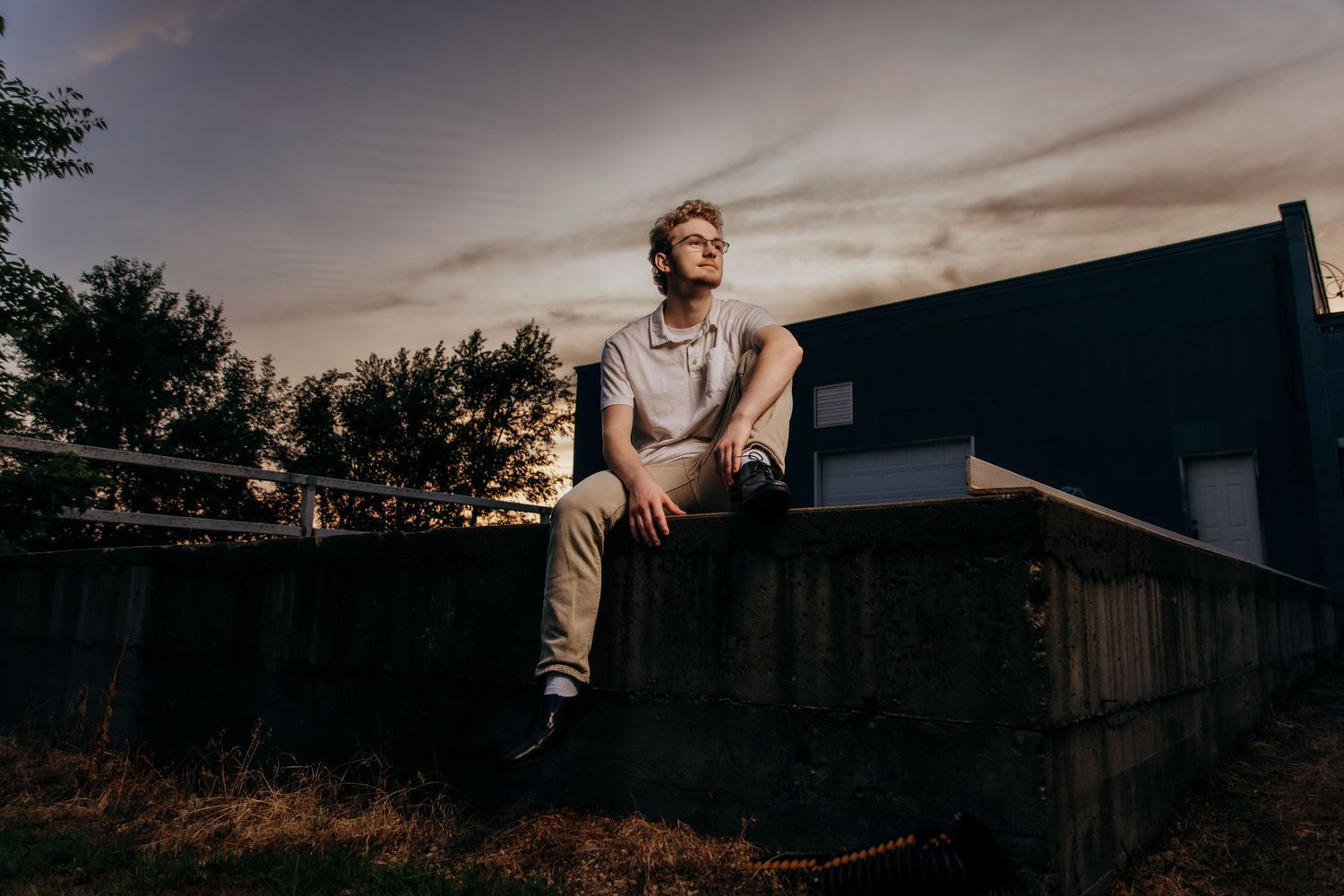 Young man with glasses sitting on a wooden fence at dusk with a dark building and cloudy sky behind him.
