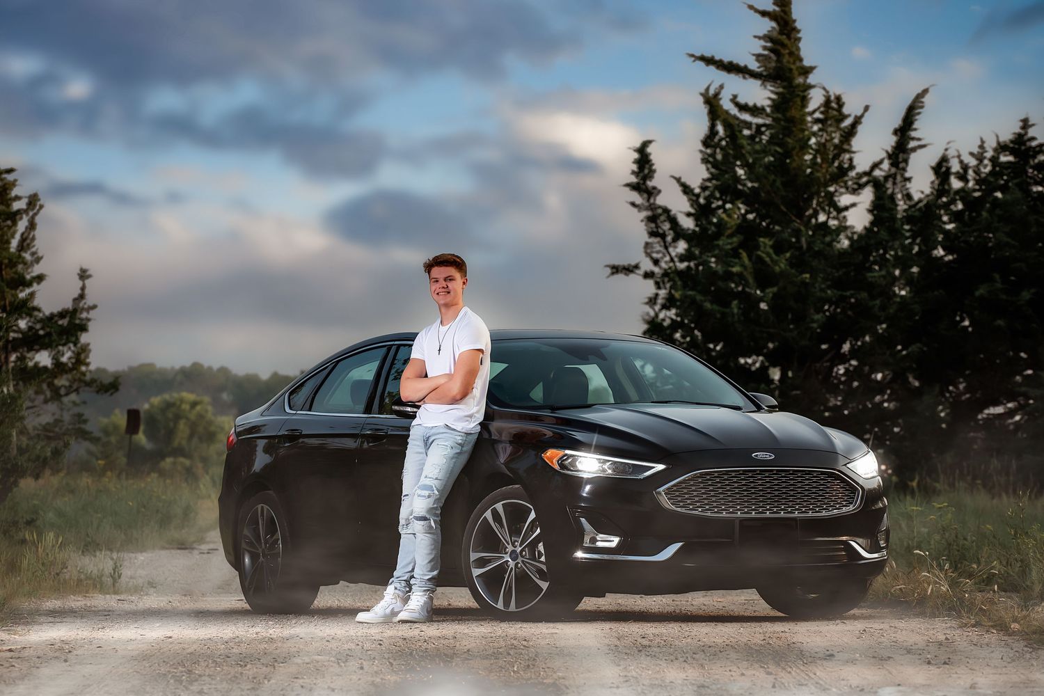 Luxury black sports car parked in scenic mountain landscape with dramatic cloudy sky at sunset.