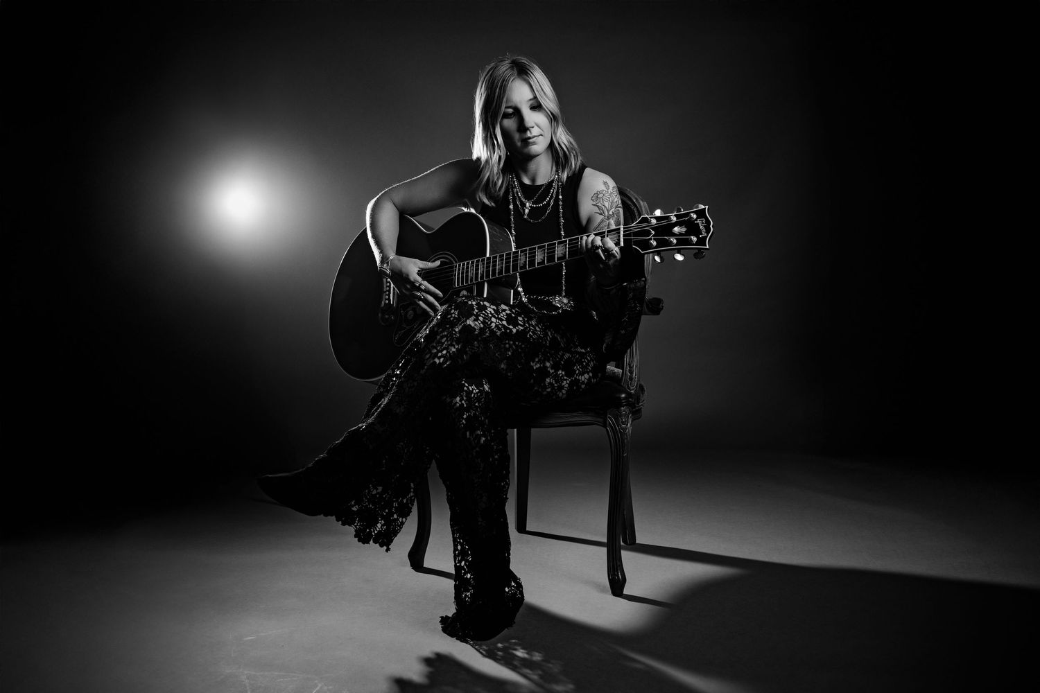 Black and white moody portrait of female guitarist seated with acoustic guitar under spotlight.
