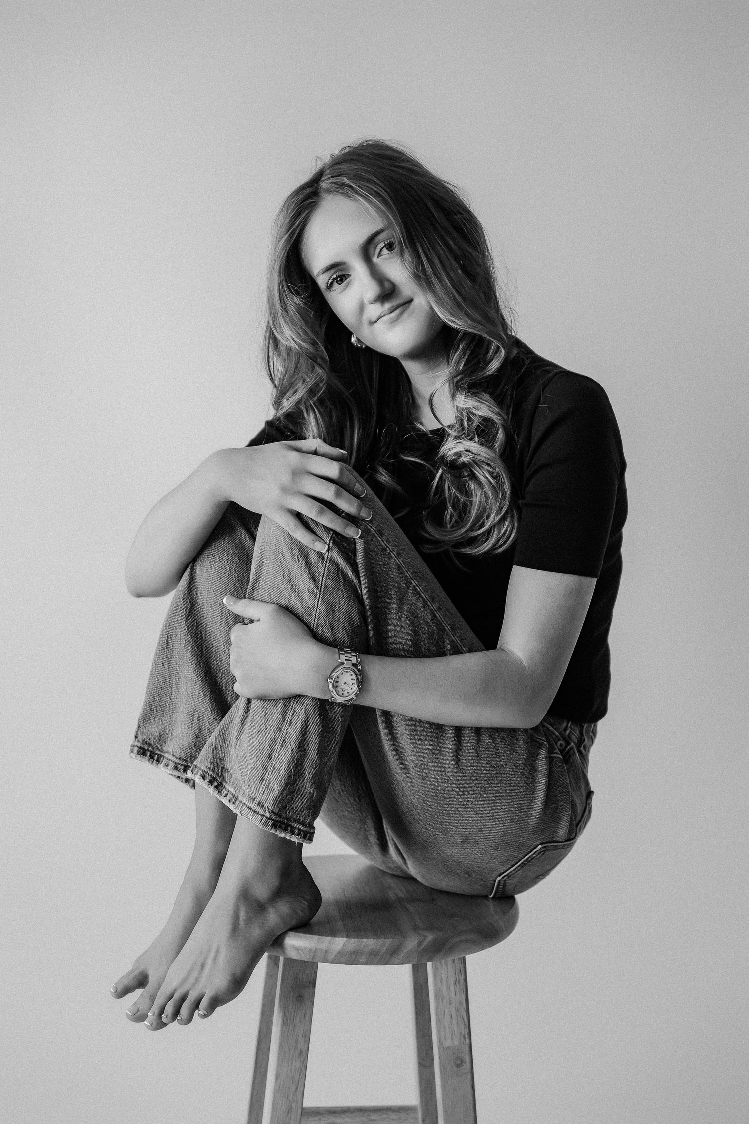 Young woman in black tee and jeans sitting on wooden stool hugging knees, black and white studio portrait.