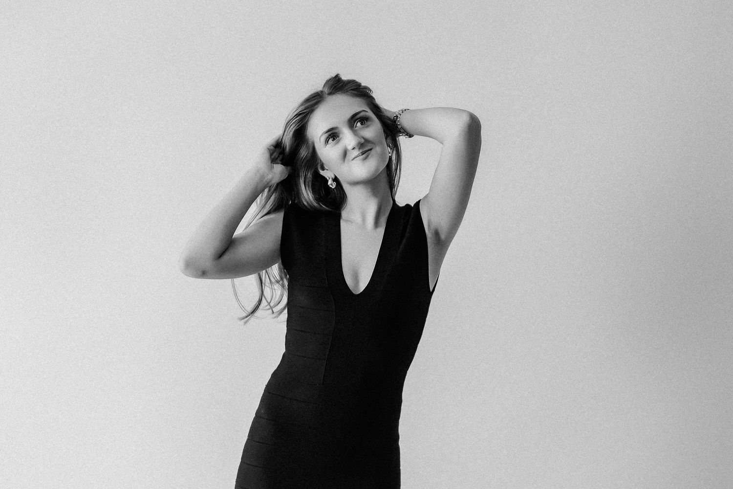 Black and white portrait of young woman in black sleeveless dress, smiling upward with hands in her hair.