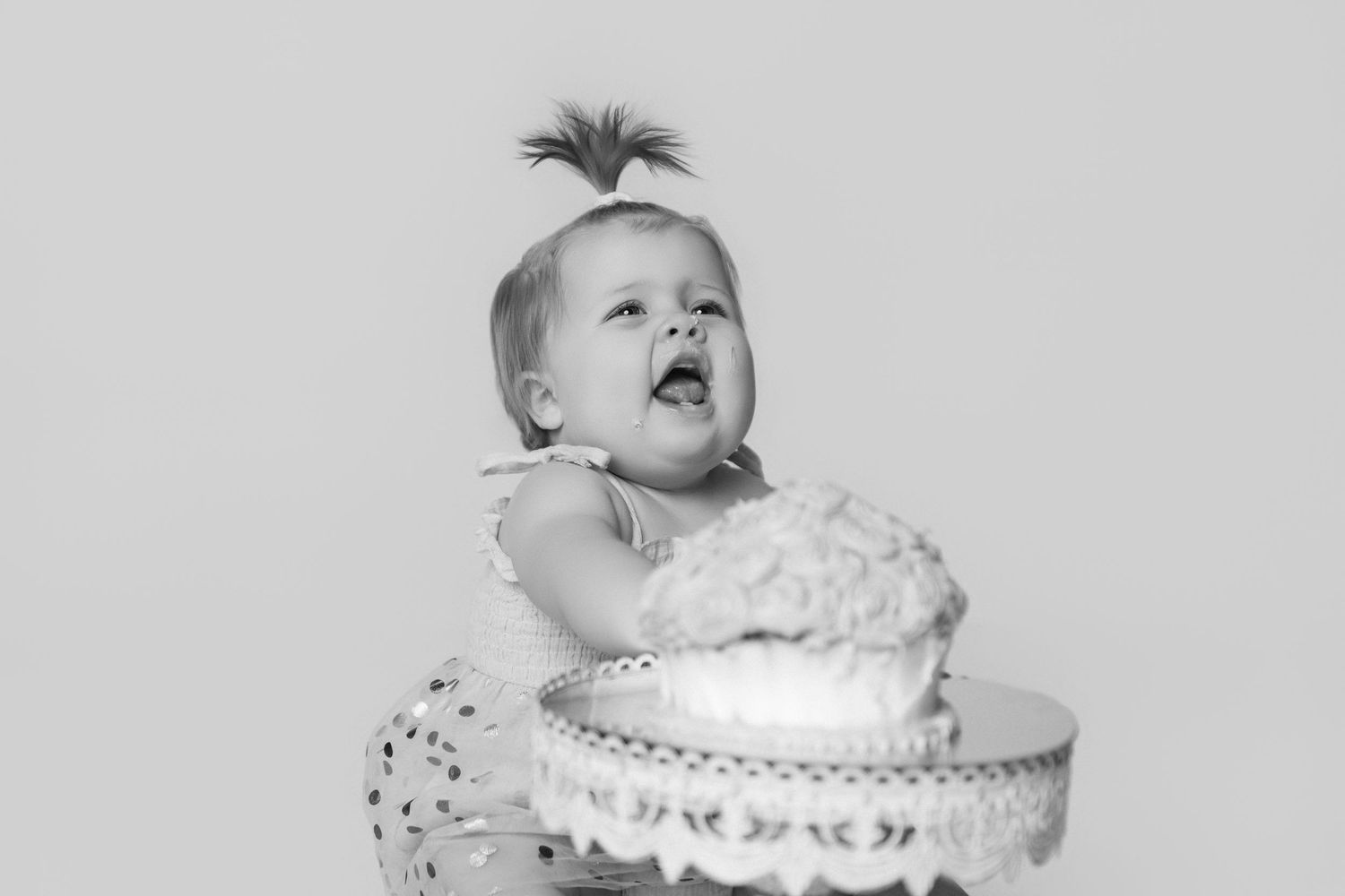 Baby in white lace outfit posing with cake in studio setting.