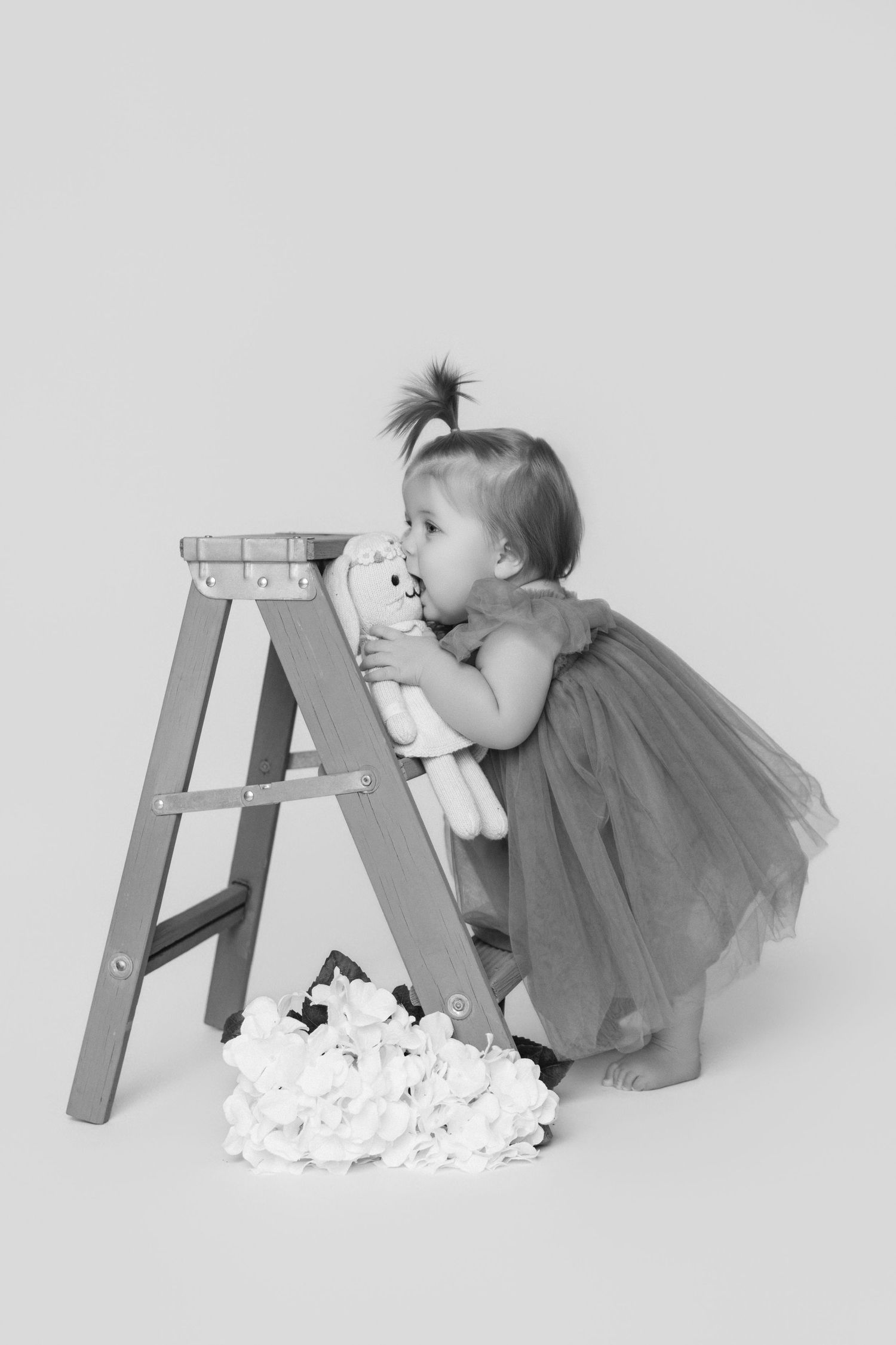 A toddler in a tulle dress peeks over a wooden step ladder with scattered white paper below.