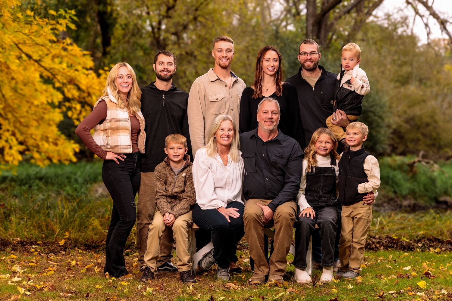 Group poses together outdoors on fallen autumn leaves with golden foliage in background during sunset photoshoot.