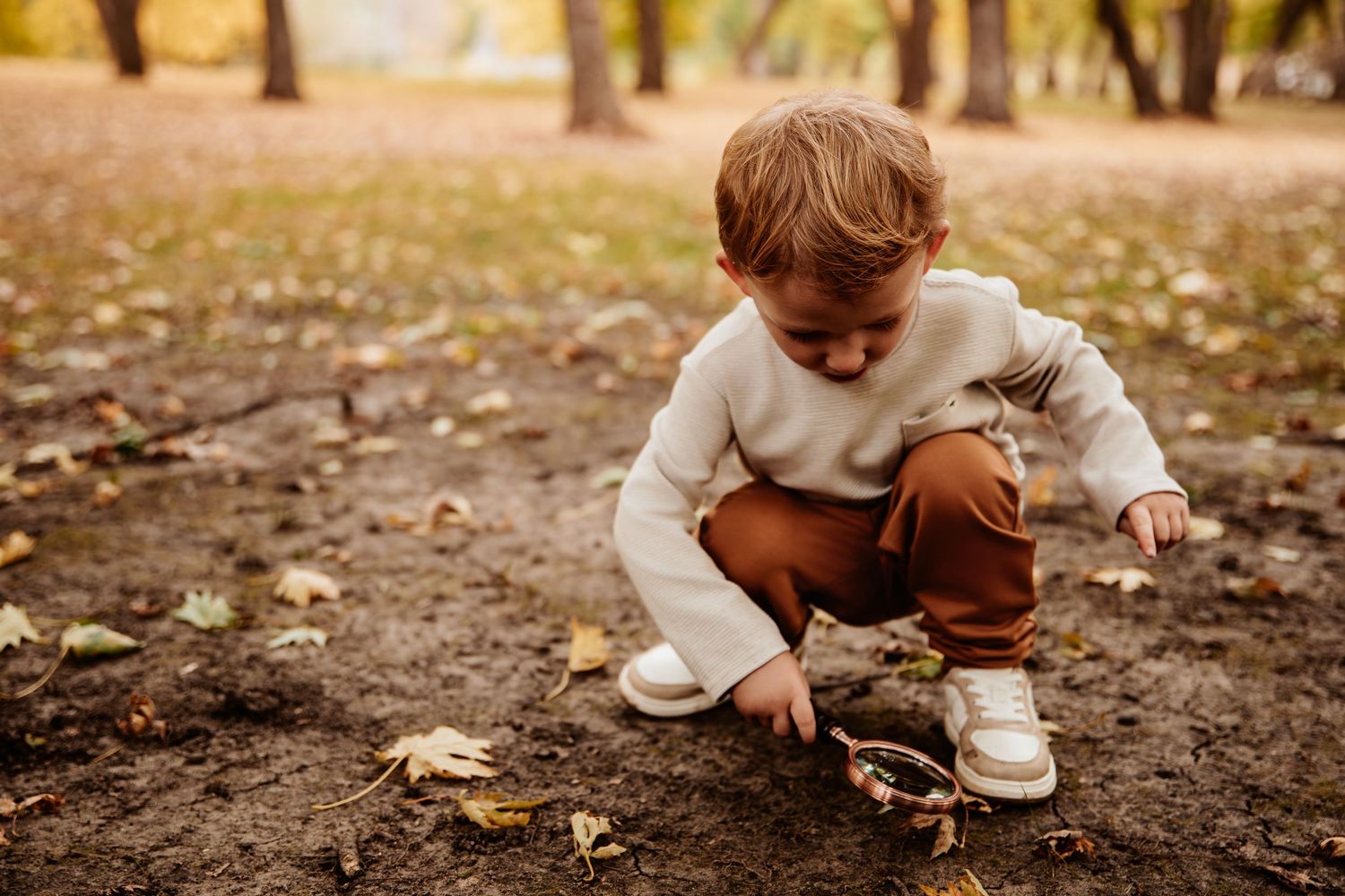 A small child crouches down to explore fallen autumn leaves in a park setting.