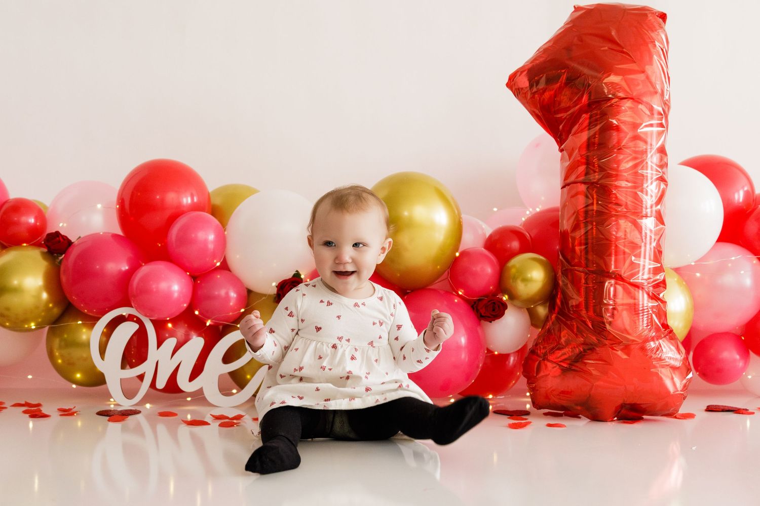 Festive first birthday setup with red and gold balloons, large number one balloon, and white 'one' sign against white backdrop.