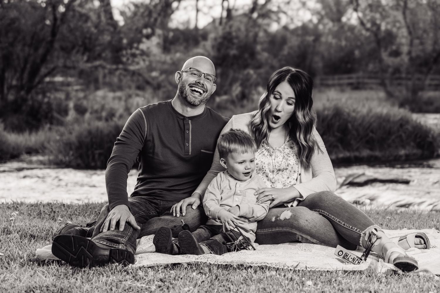 Black and white photo of a laughing family with a toddler sitting on a blanket in a park.