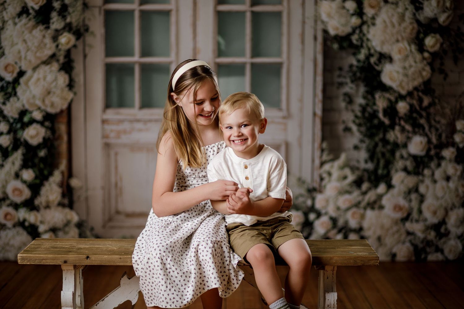 A young girl in a floral dress sits with a toddler boy on a bench surrounded by white roses.