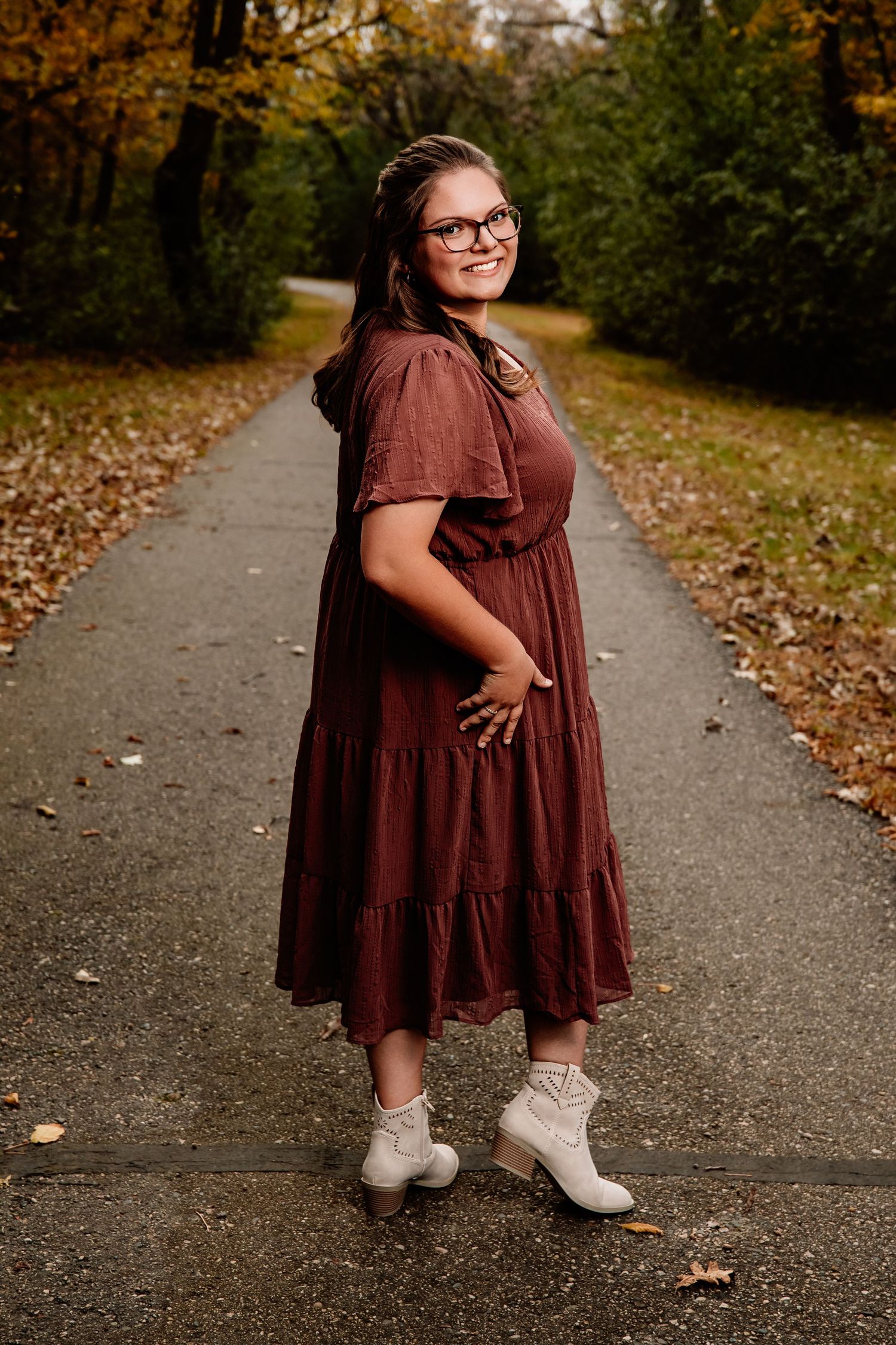 A person in a burgundy dress and white boots poses on a leaf-scattered path during autumn.