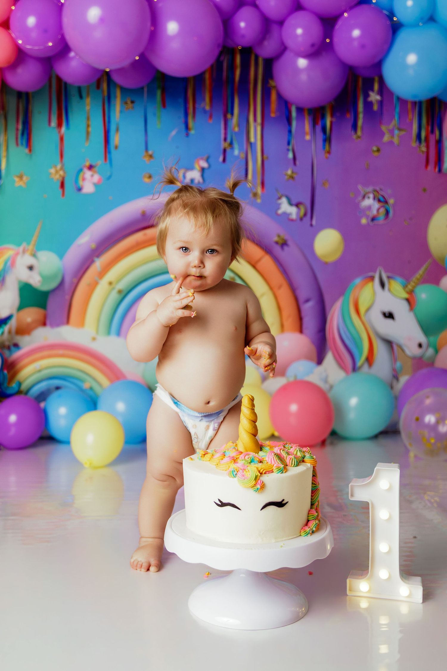 Baby girl celebrates first birthday with unicorn cake and colorful balloon backdrop.
