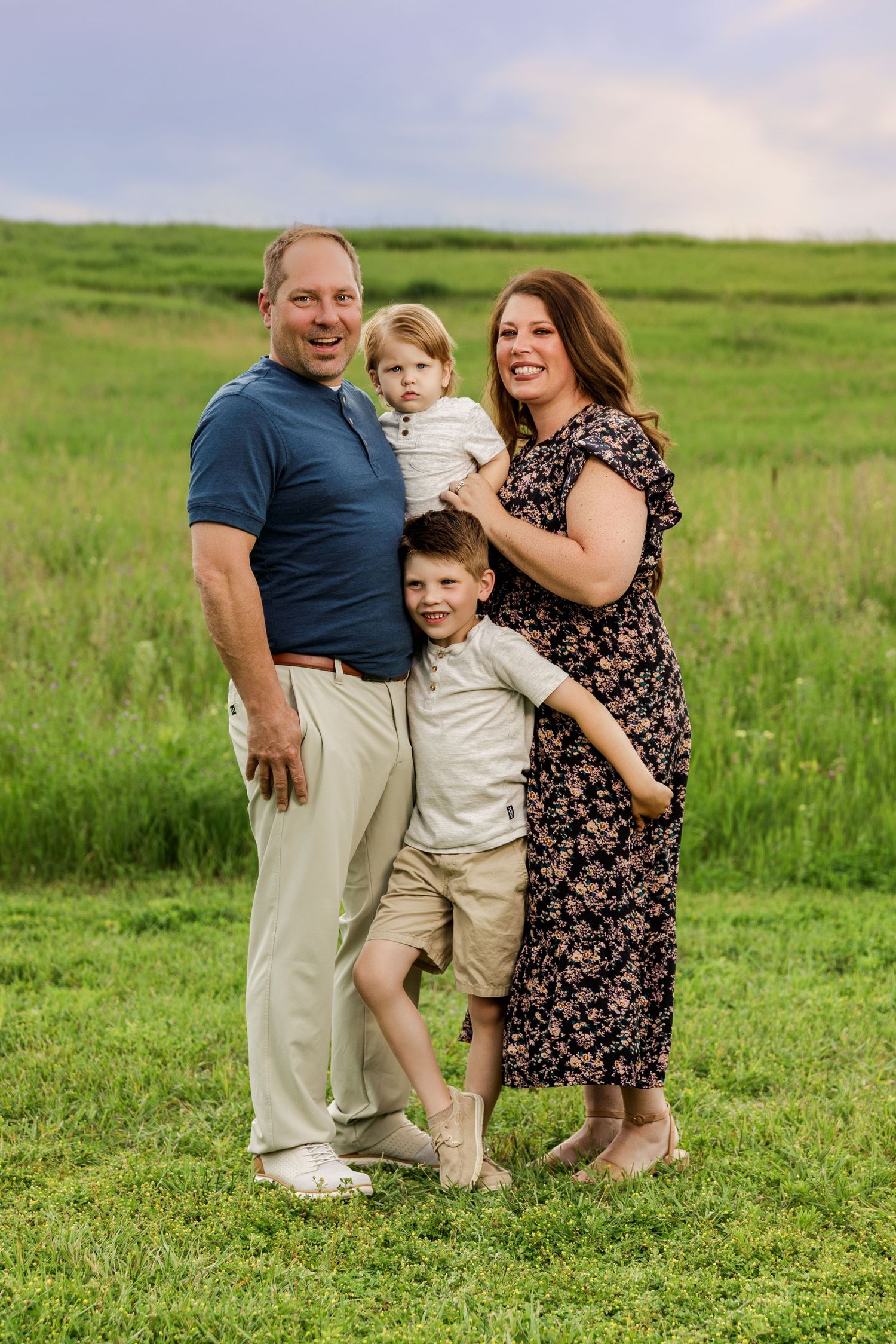 Family of four posing outdoors in a green field, parents holding two young boys, smiling for a portrait photo.