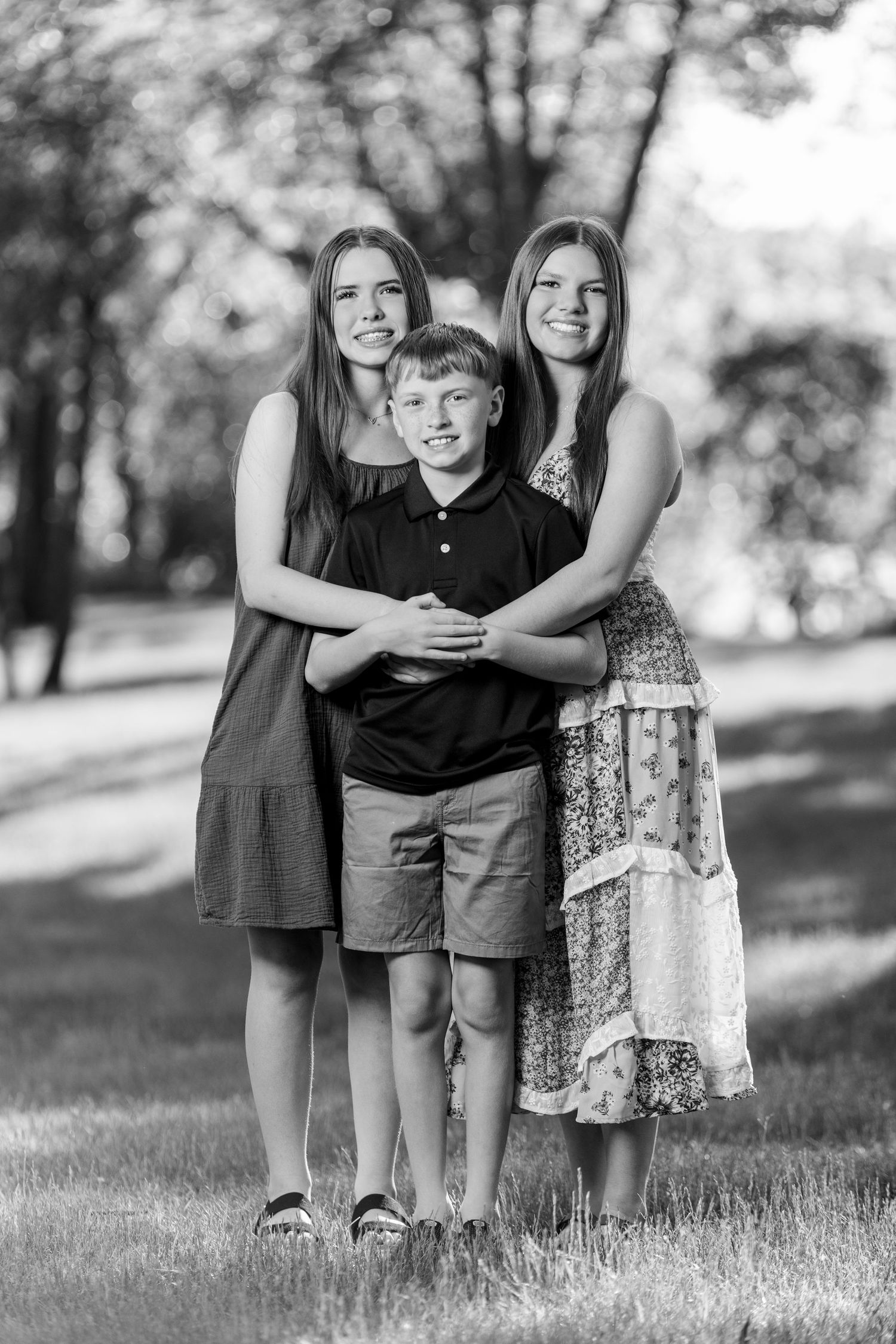 Black and white photo of three siblings posing outdoors in a park, two teenage girls hugging a young boy.