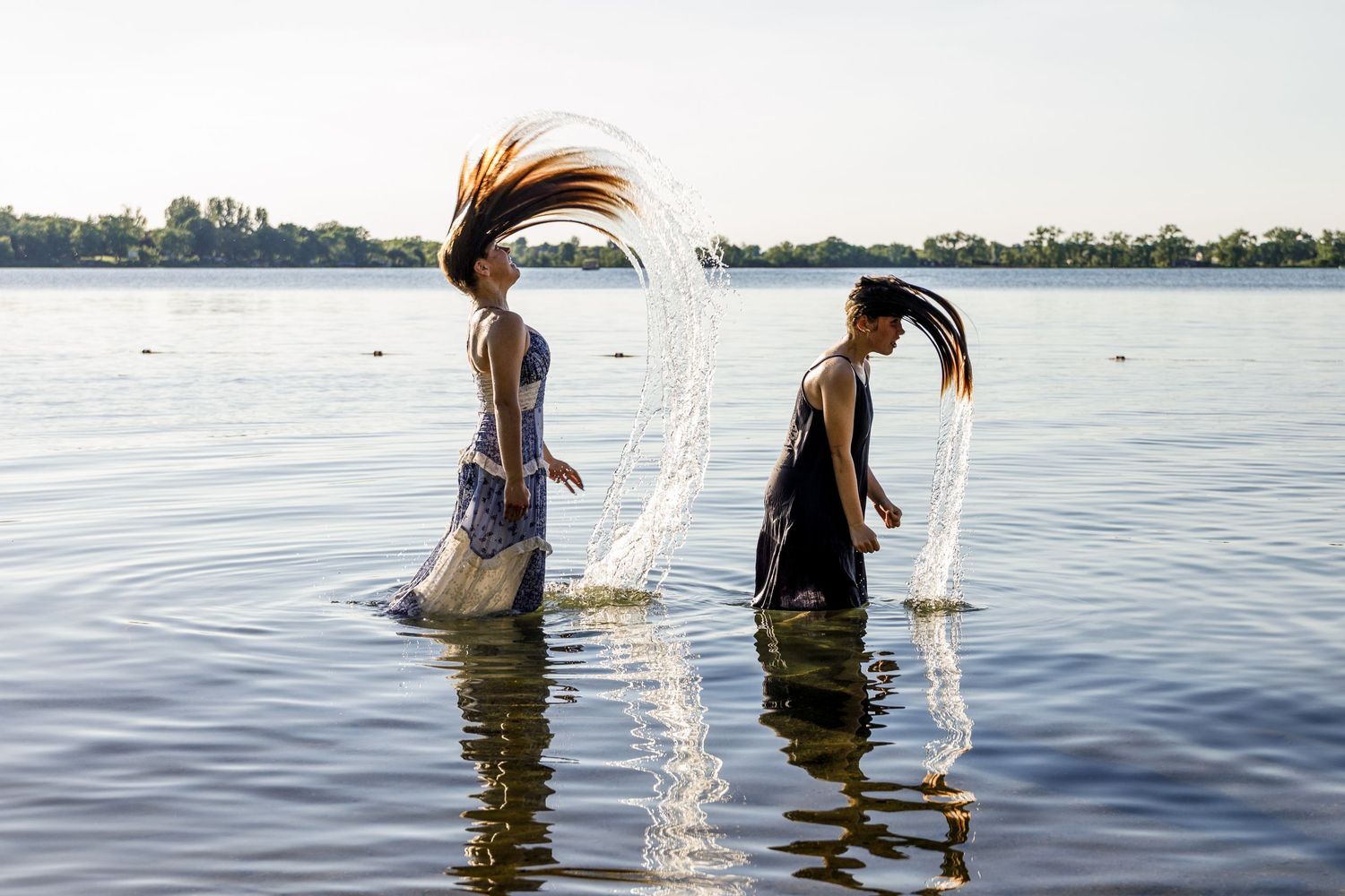 Two women in dresses flipping their wet hair in a lake creating water arcs in the sunlight.