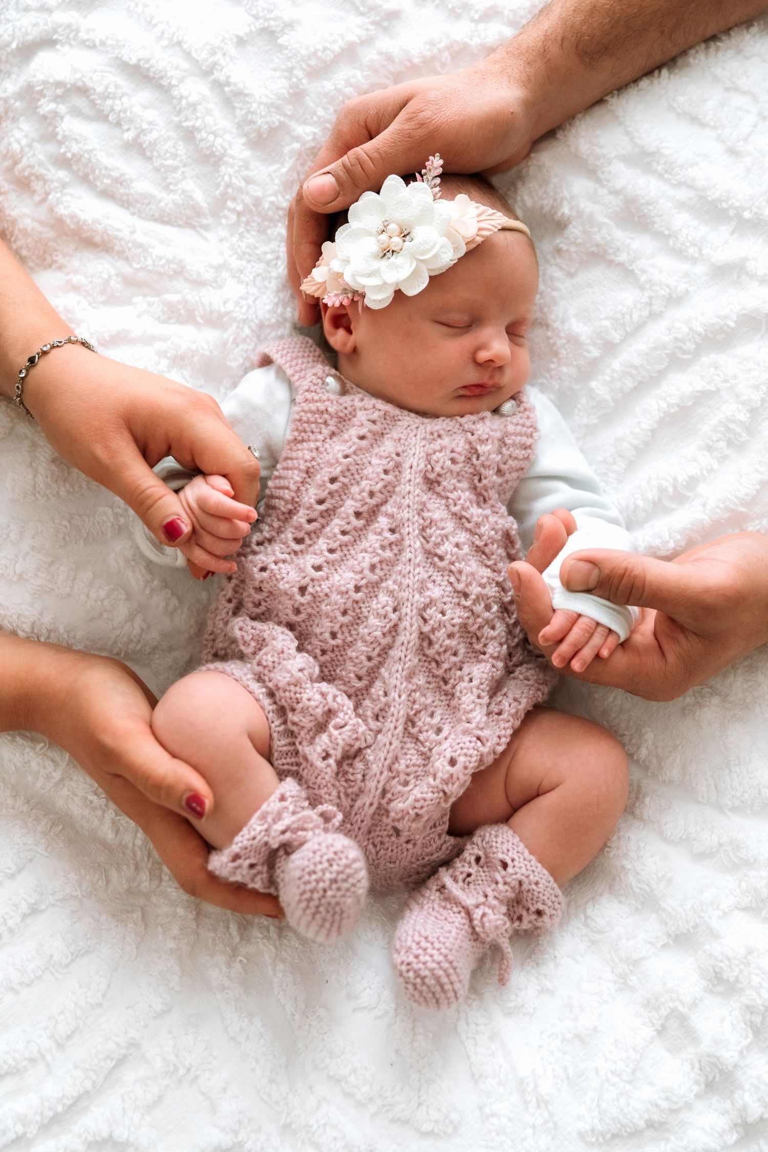 A sleeping newborn wearing a delicate pink lace romper lies on white textured bedding while hands gently touch their feet.