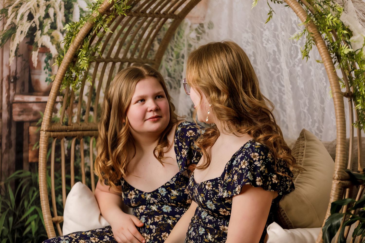 Two young girls in matching floral dresses sharing a moment in a decorative wicker swing chair.