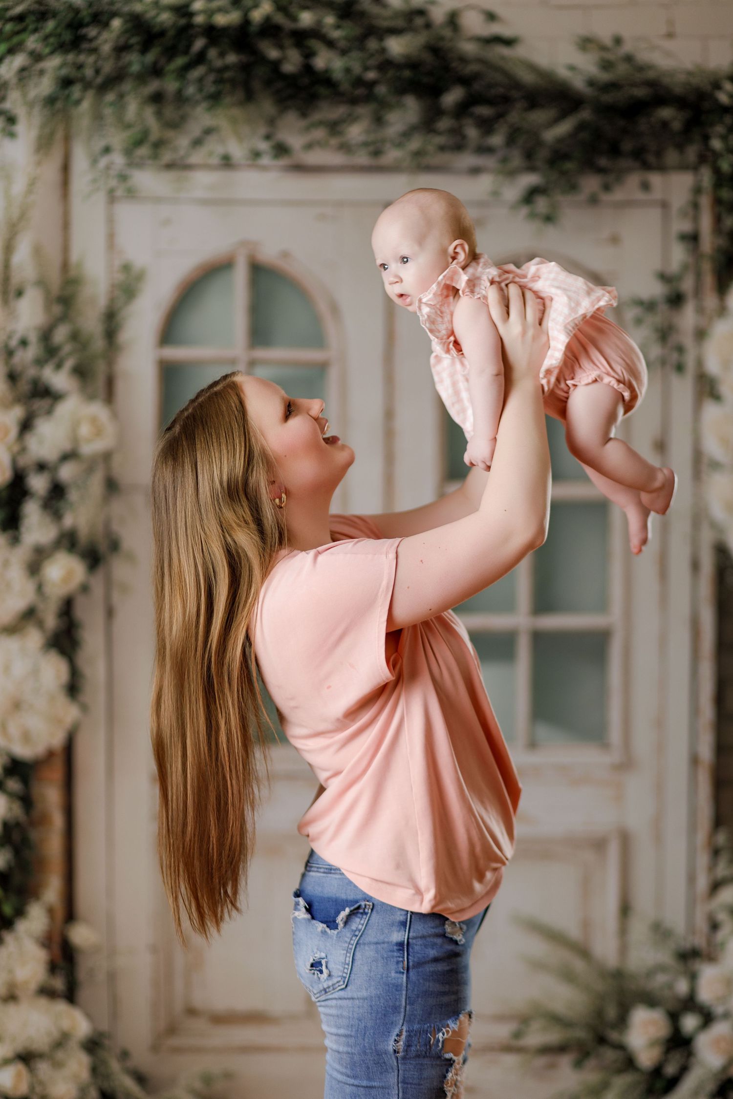 Young mother in pink top lifts smiling baby girl in pink dress against floral decorated backdrop.