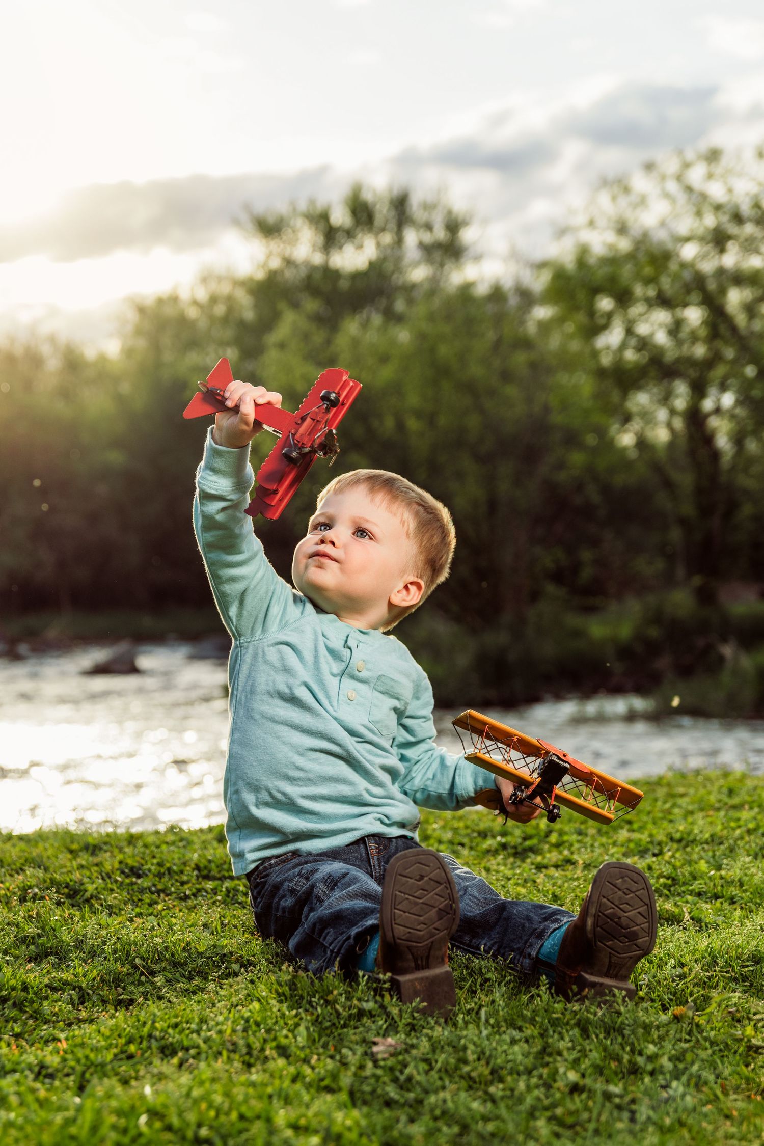 Young boy sitting on grass holding red toy airplanes up in the air outdoors near a river.