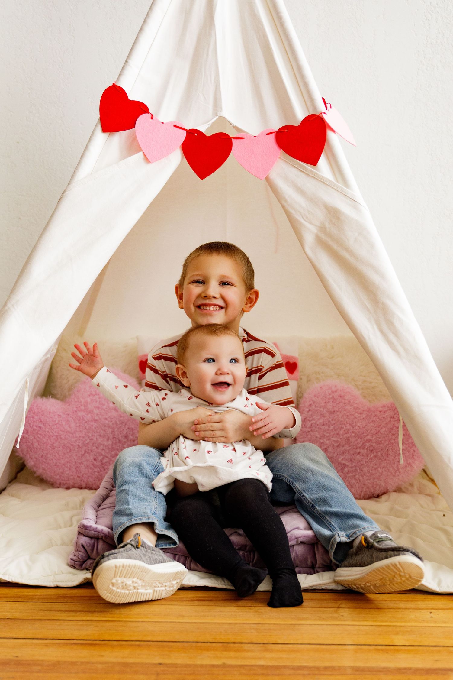 Two small children sit together and laugh inside a white teepee tent decorated with red and pink heart garlands.