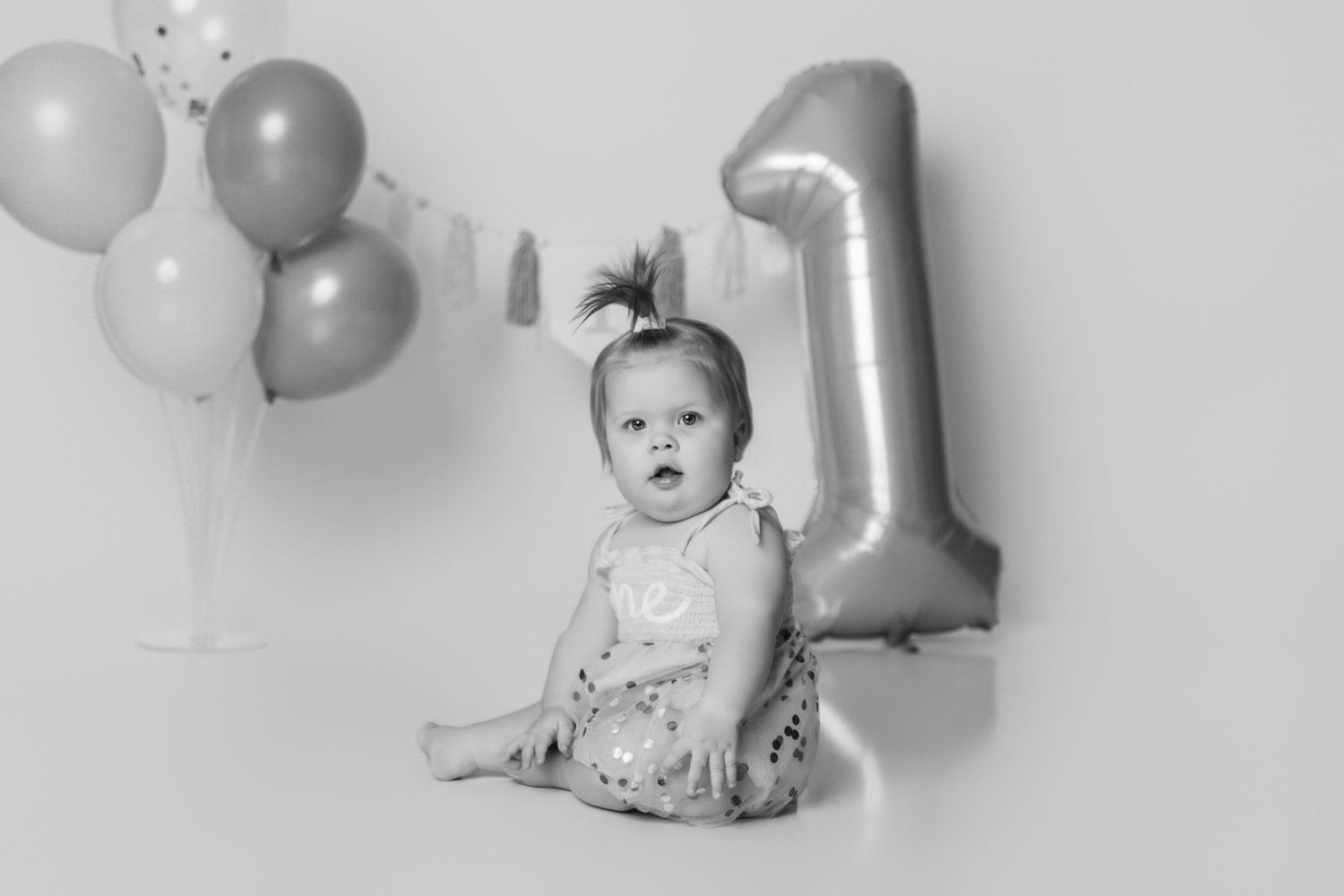 A baby sits on the floor surrounded by balloons and a large silver number one balloon.