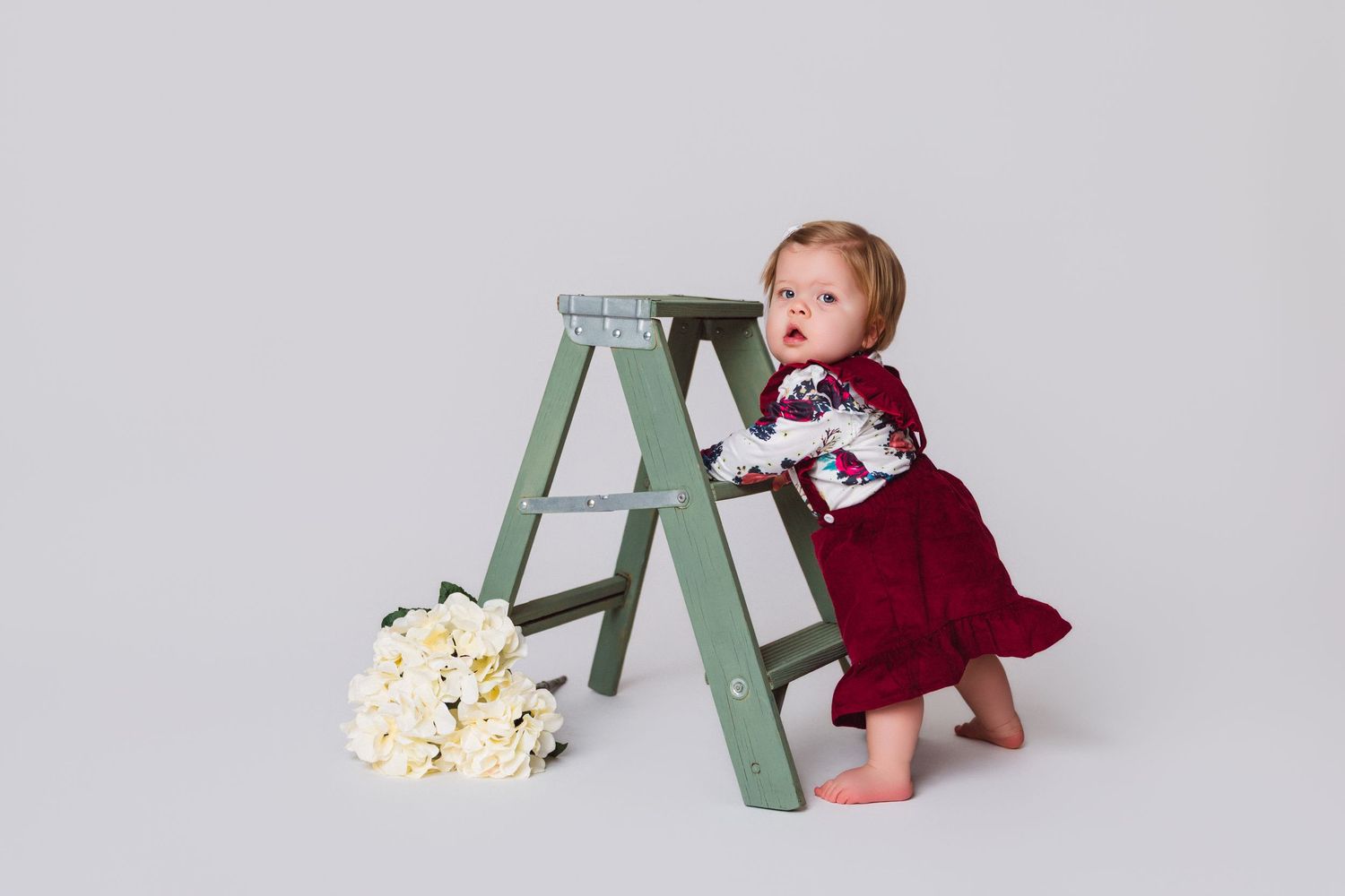 A baby in a red dress sitting near a mint green ladder with scattered popcorn on the floor.