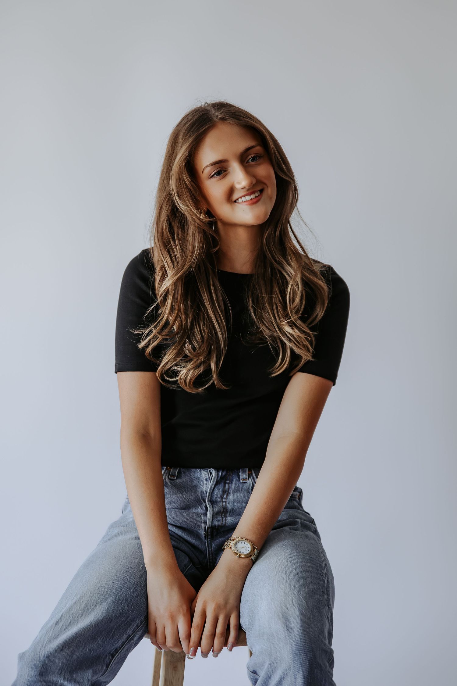Young woman with long wavy hair smiles warmly while seated on stool, wearing black tee, jeans, and gold watch.