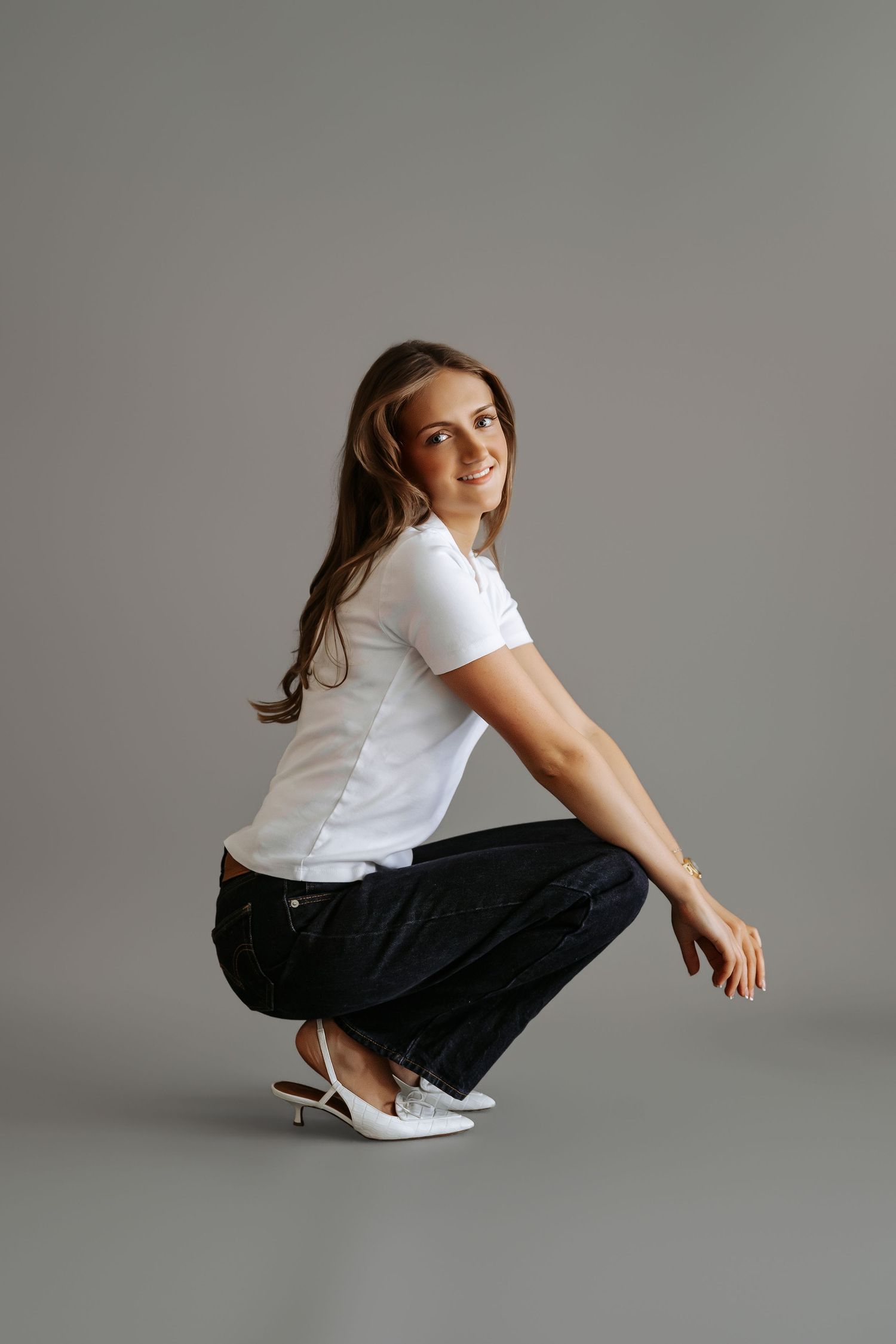 Smiling young woman in white t-shirt and dark jeans crouches gracefully in white heels against gray background.