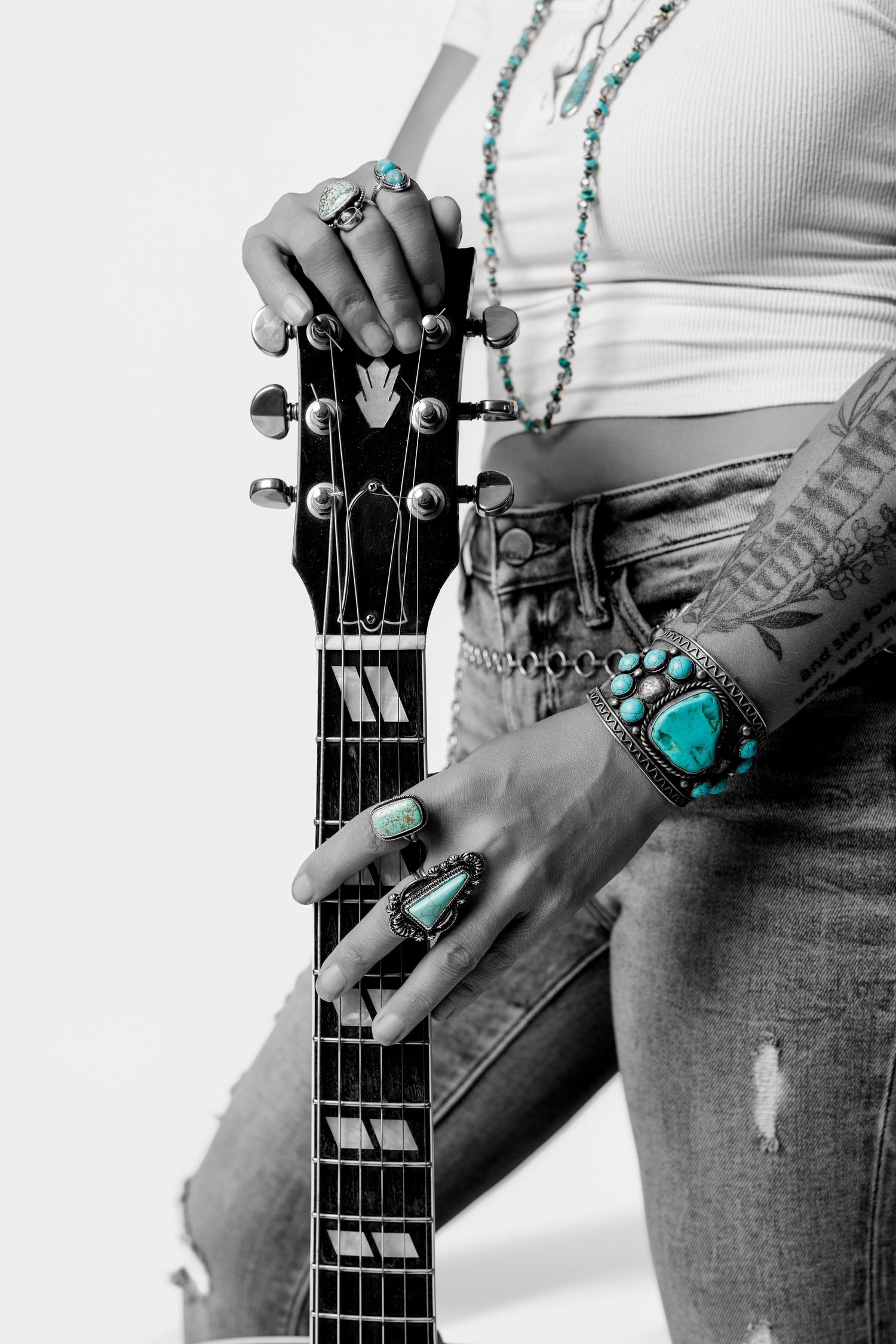 Close-up of tattooed hands holding guitar neck adorned with turquoise rings and bracelet in selective color.