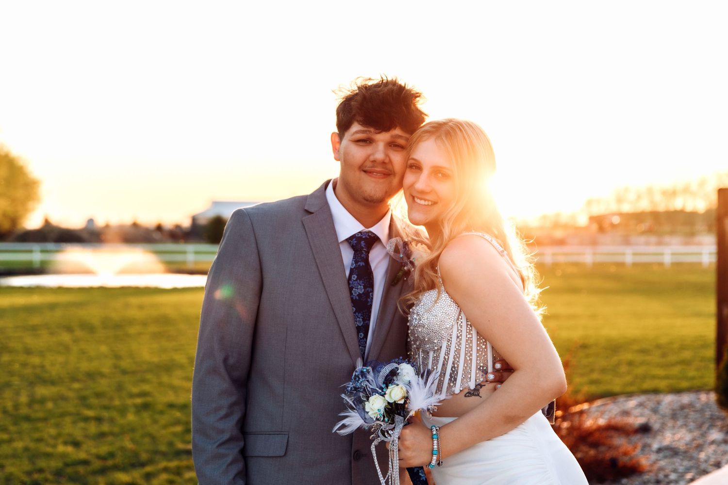 A newlywed couple embraces during golden hour sunset on a grassy field with a bouquet of blue flowers.