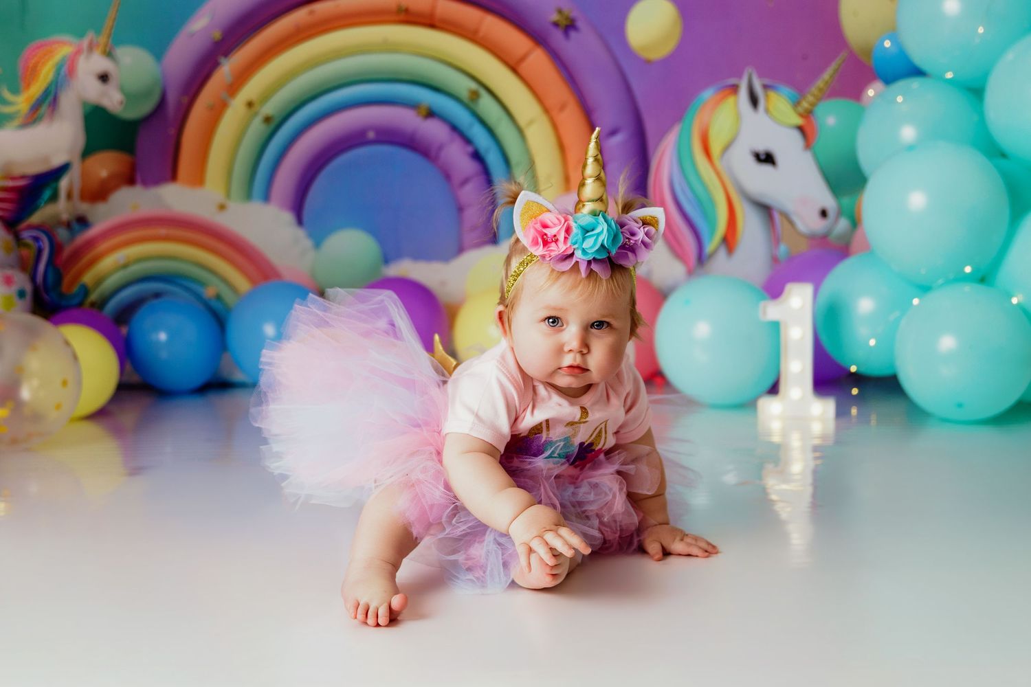 Baby girl in rainbow tutu and unicorn headband sits before colorful balloon backdrop at 1st birthday party.