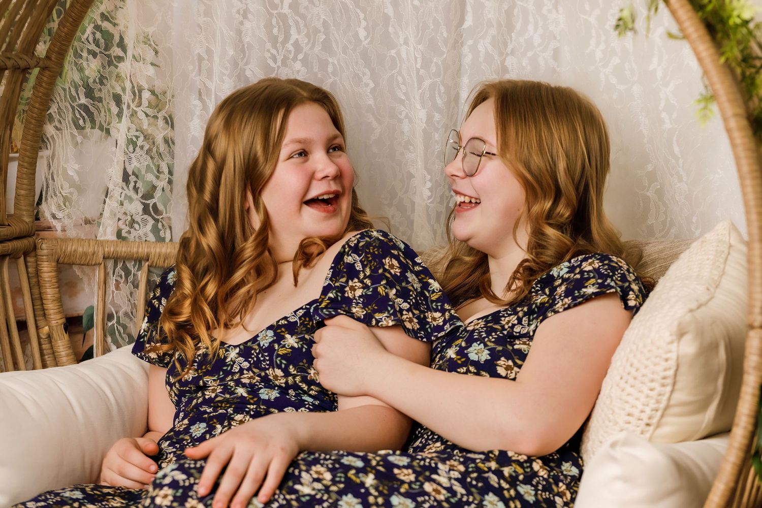 Two young girls in matching floral dresses laughing together while sitting in a wicker chair.