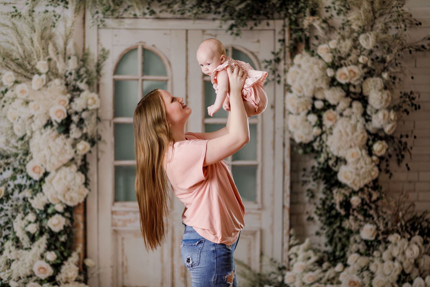 A mother joyfully lifts her baby girl in a floral studio setting with white roses and greenery.