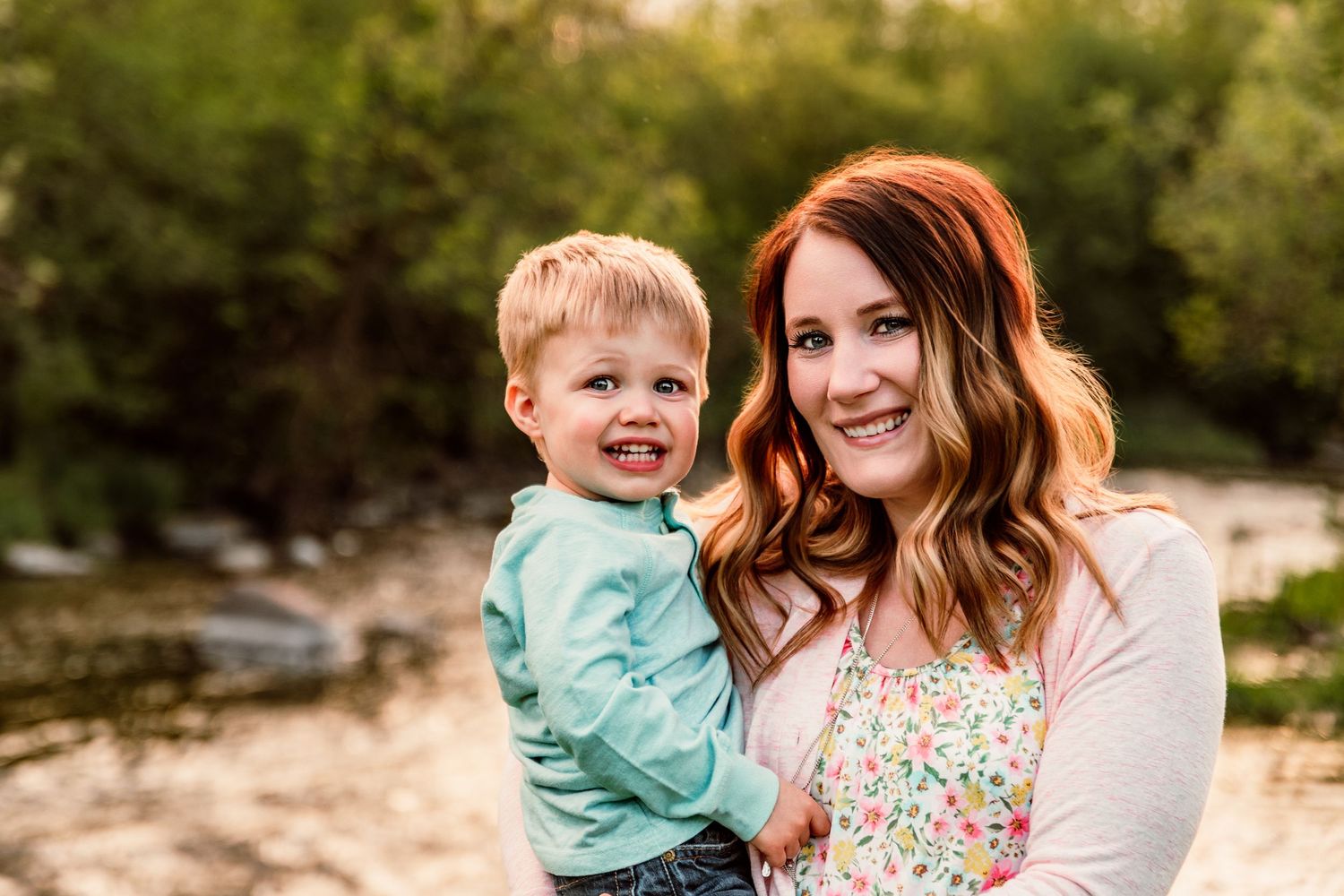 A smiling mother with auburn hair holds her happy toddler boy outdoors near a creek with lush greenery.