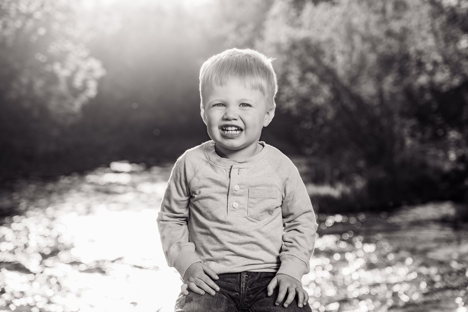 Black and white portrait of a smiling toddler boy standing outdoors near a sunlit river.