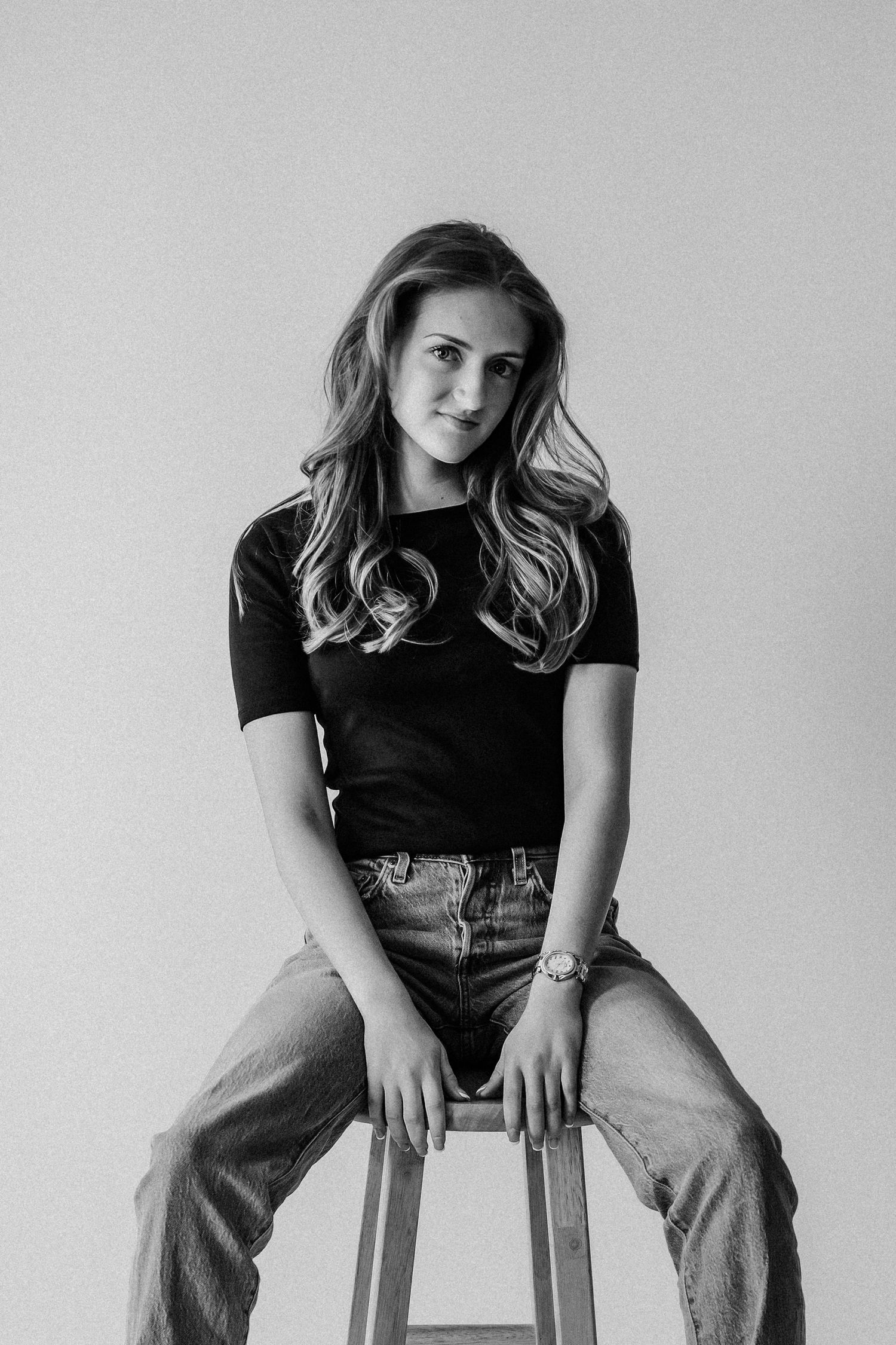 Young woman with wavy hair sits on a stool in black tee and jeans, black and white studio portrait.