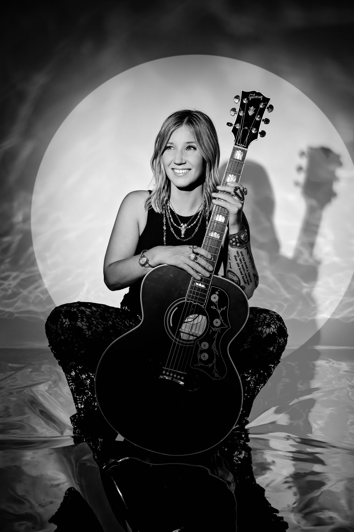 Smiling female musician poses with black Gibson guitar in black and white photo with circular light backdrop.