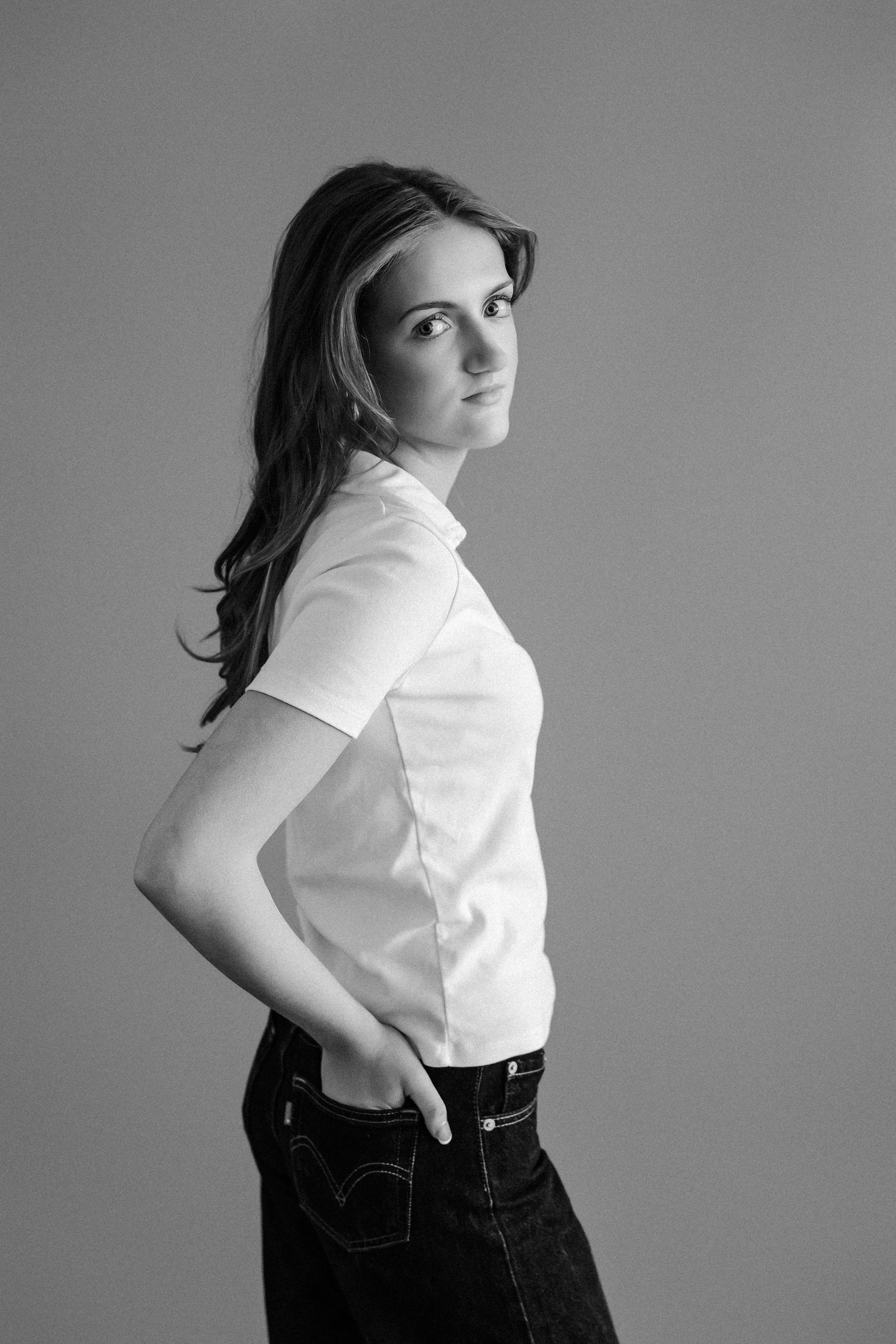 Young woman in white shirt and jeans posing sideways, black and white studio portrait.