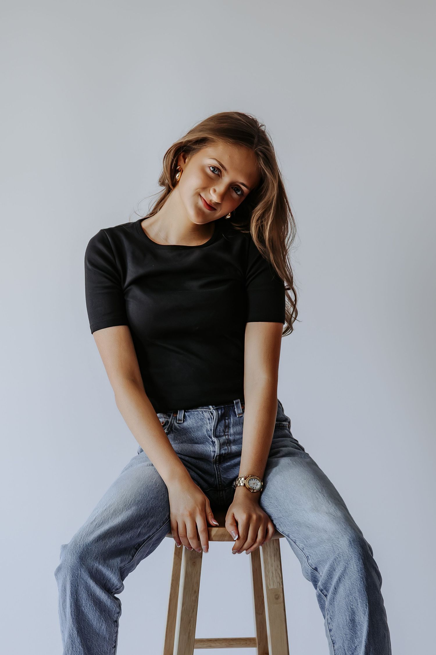 Young woman in black t-shirt and jeans sitting on wooden stool against white background, smiling at camera.