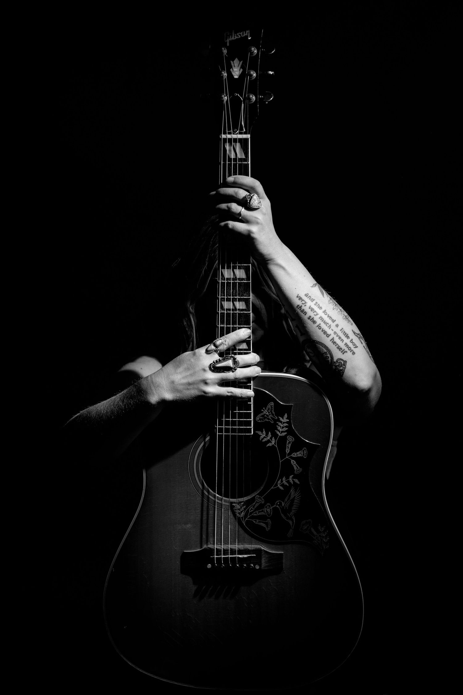 Black and white photo of tattooed hands holding an acoustic guitar against a dark background.