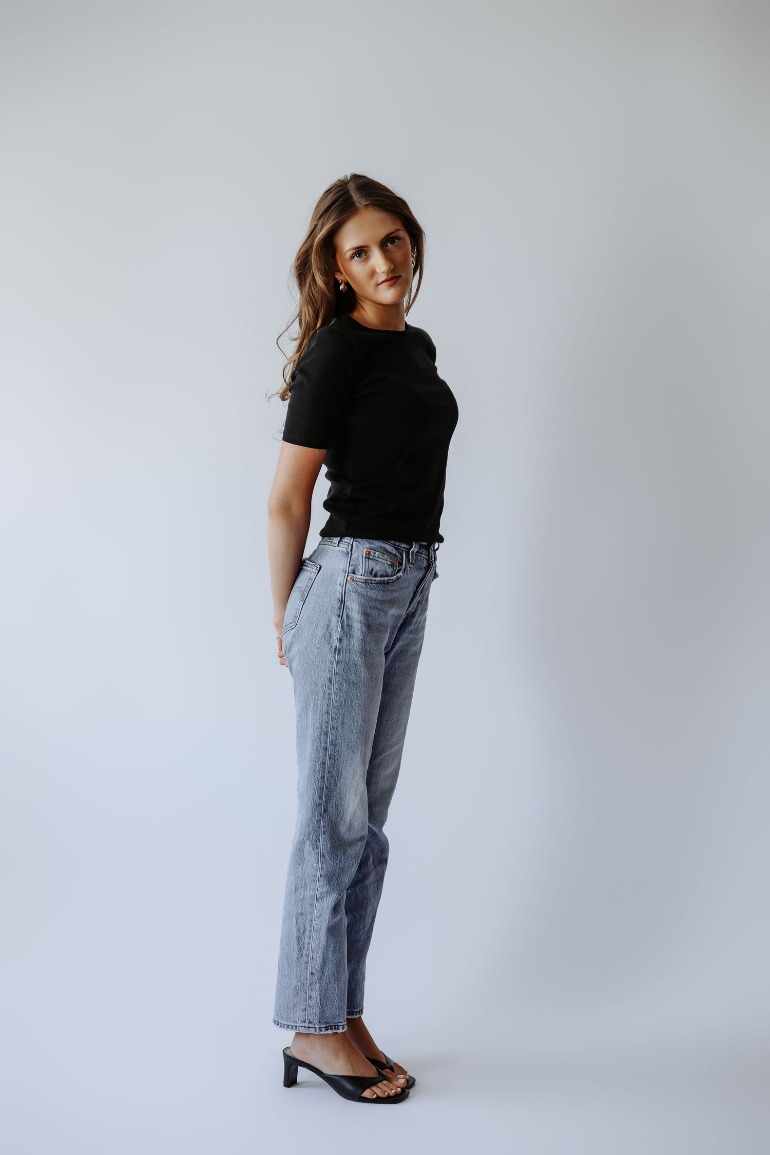 Young woman in black t-shirt, light jeans, and heeled sandals poses sideways in minimalist studio setting.