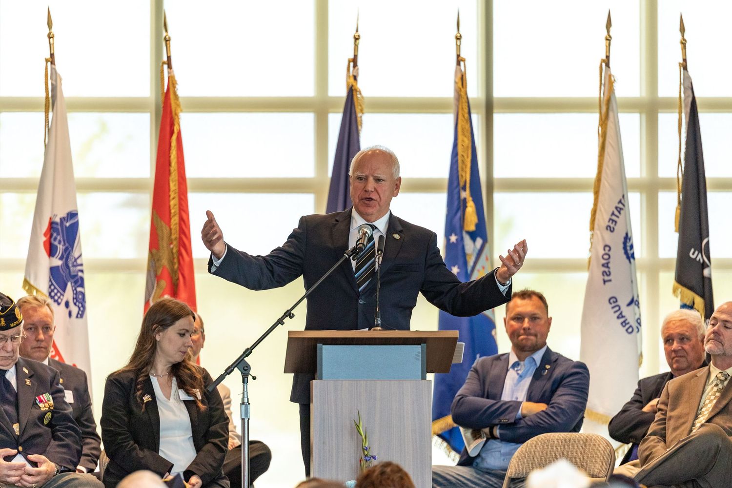 Politician speaks passionately at podium before audience with military branch flags displayed behind him.