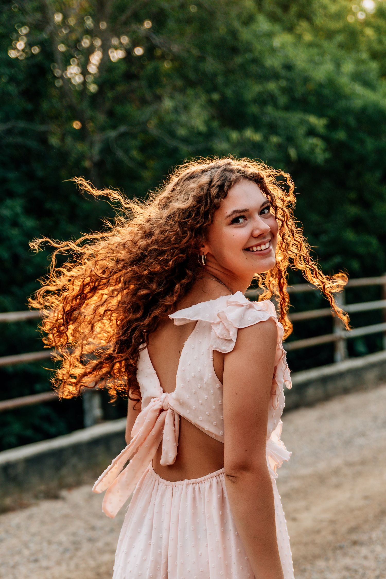 Young woman with curly hair smiling over shoulder in pink backless dress outdoors at golden hour.