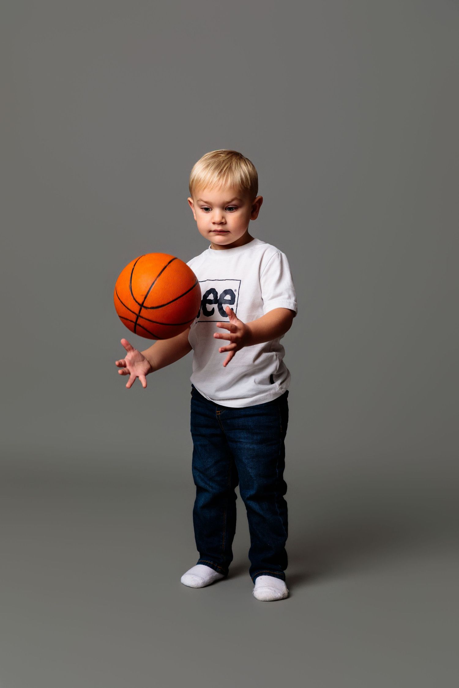Small child in white Lee shirt holding orange basketball while standing against gray backdrop.